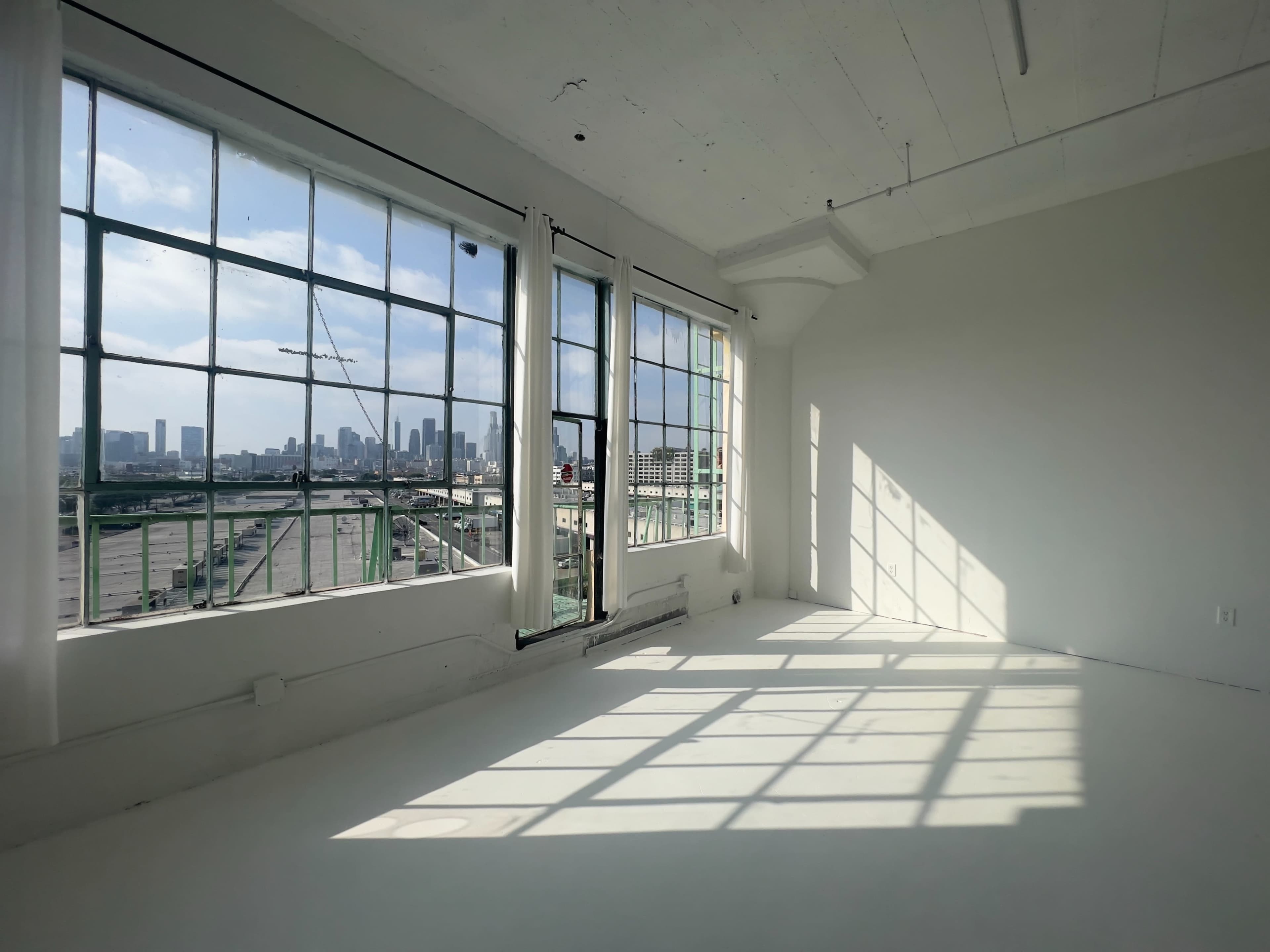 A bright, empty room with large windows offers a view of a city skyline and casts long shadows on the white floor.