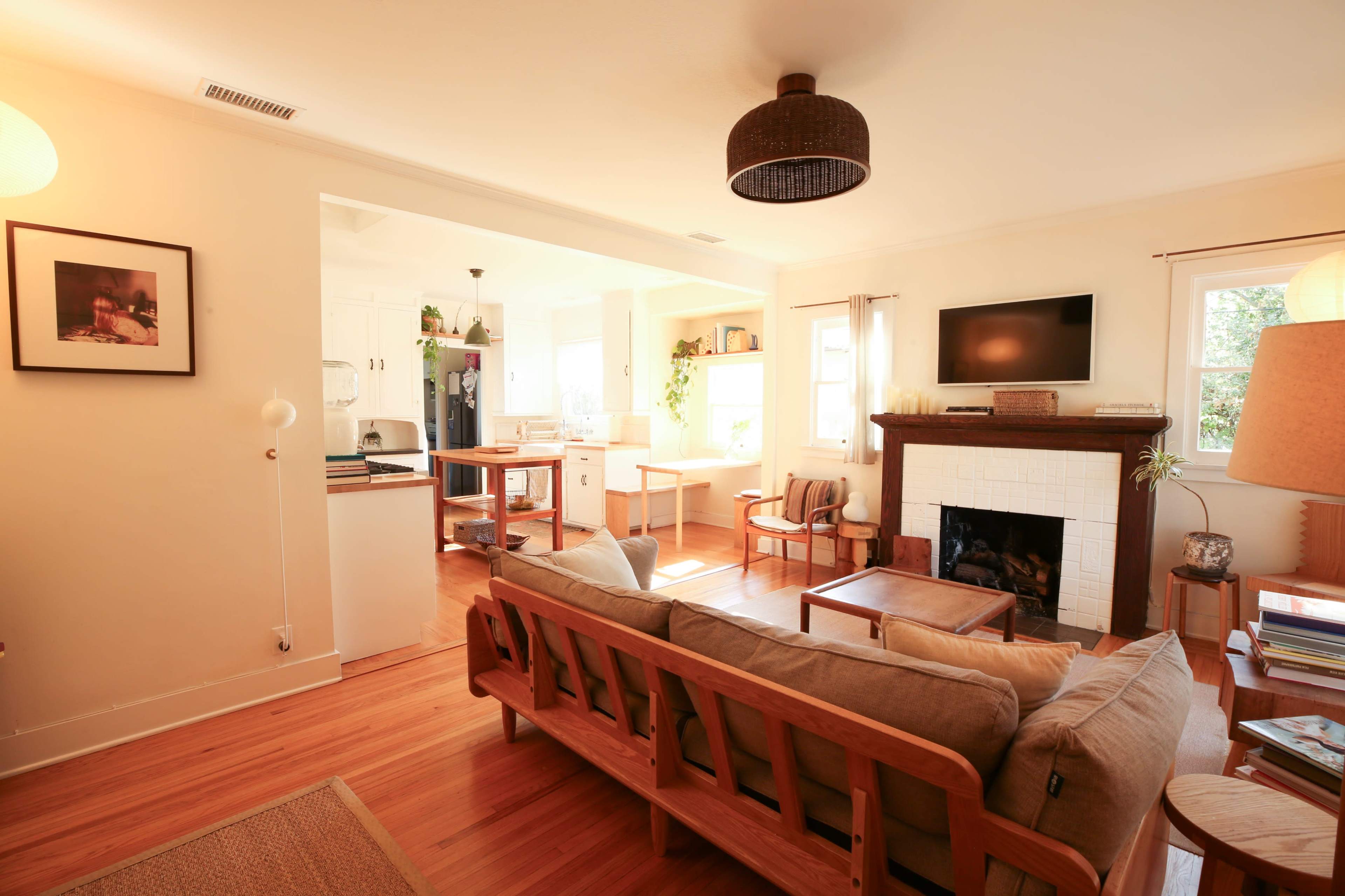 The image shows a cozy living room featuring a wooden couch, a fireplace, and an open kitchen area with natural light streaming through the windows.