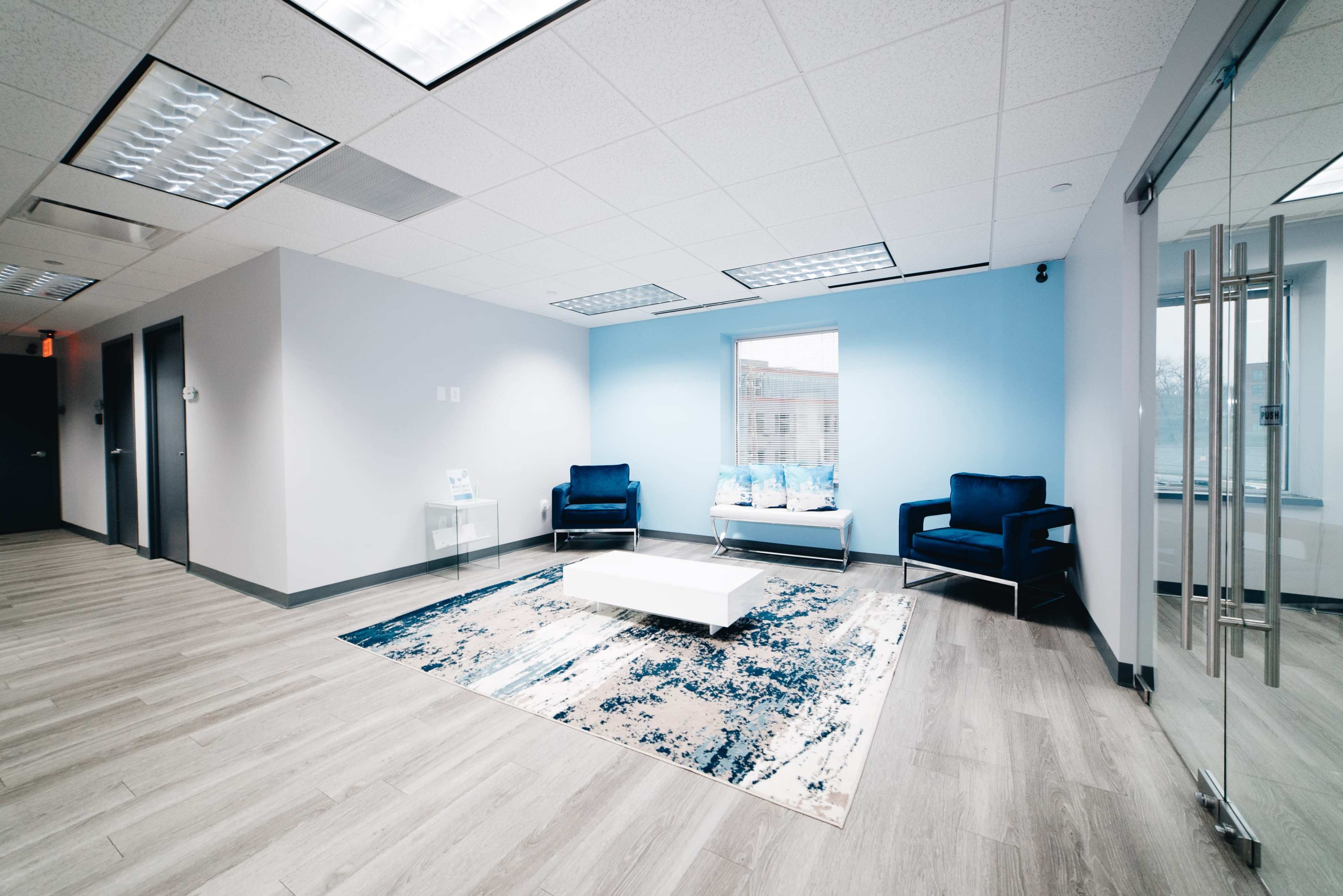 A modern office waiting area with two blue chairs, a white coffee table, and light blue wall accents, featuring a rug on a wood floor.