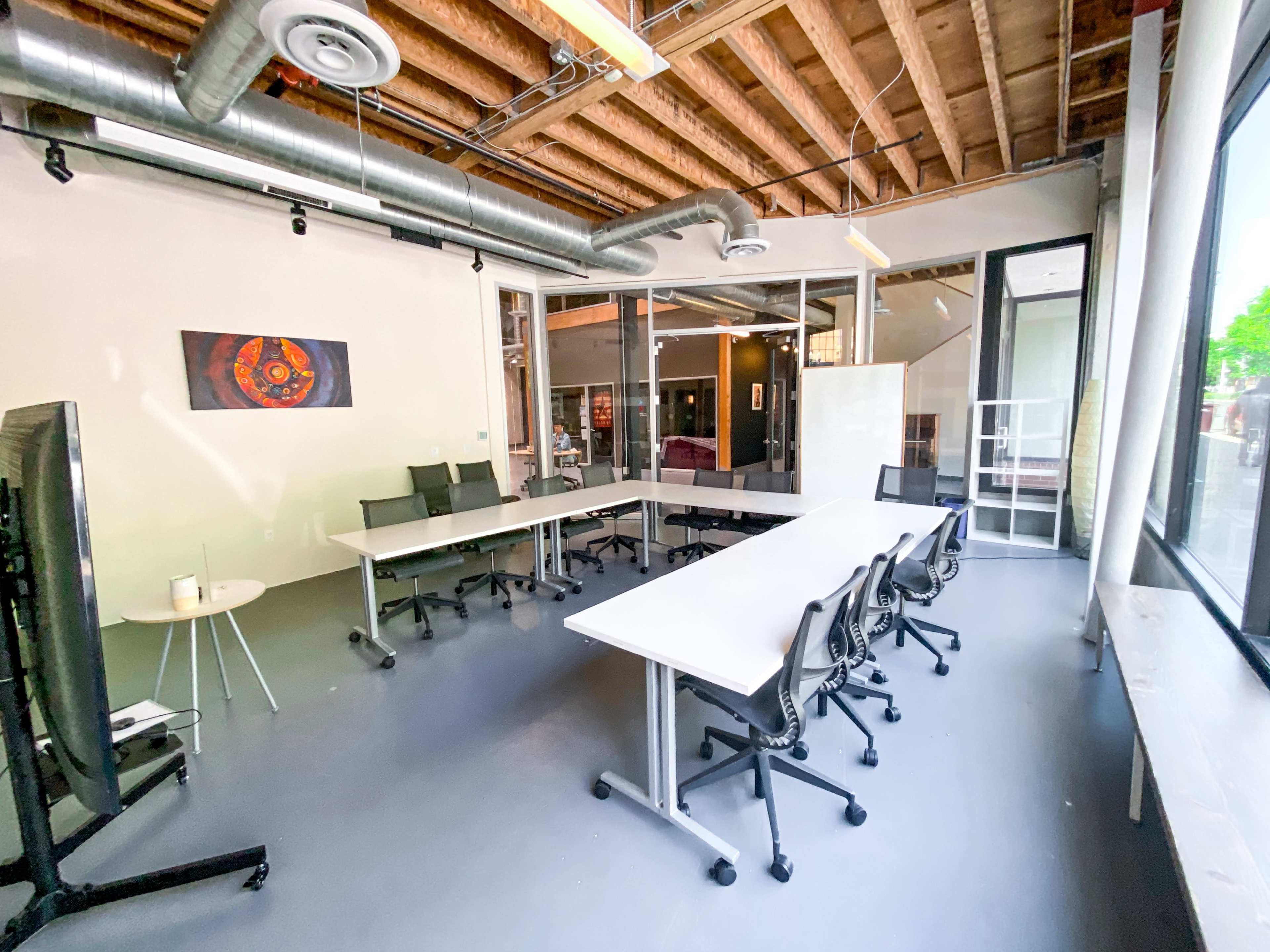 A modern conference room features a large table surrounded by chairs, with windows allowing natural light and a piece of artwork on the wall.