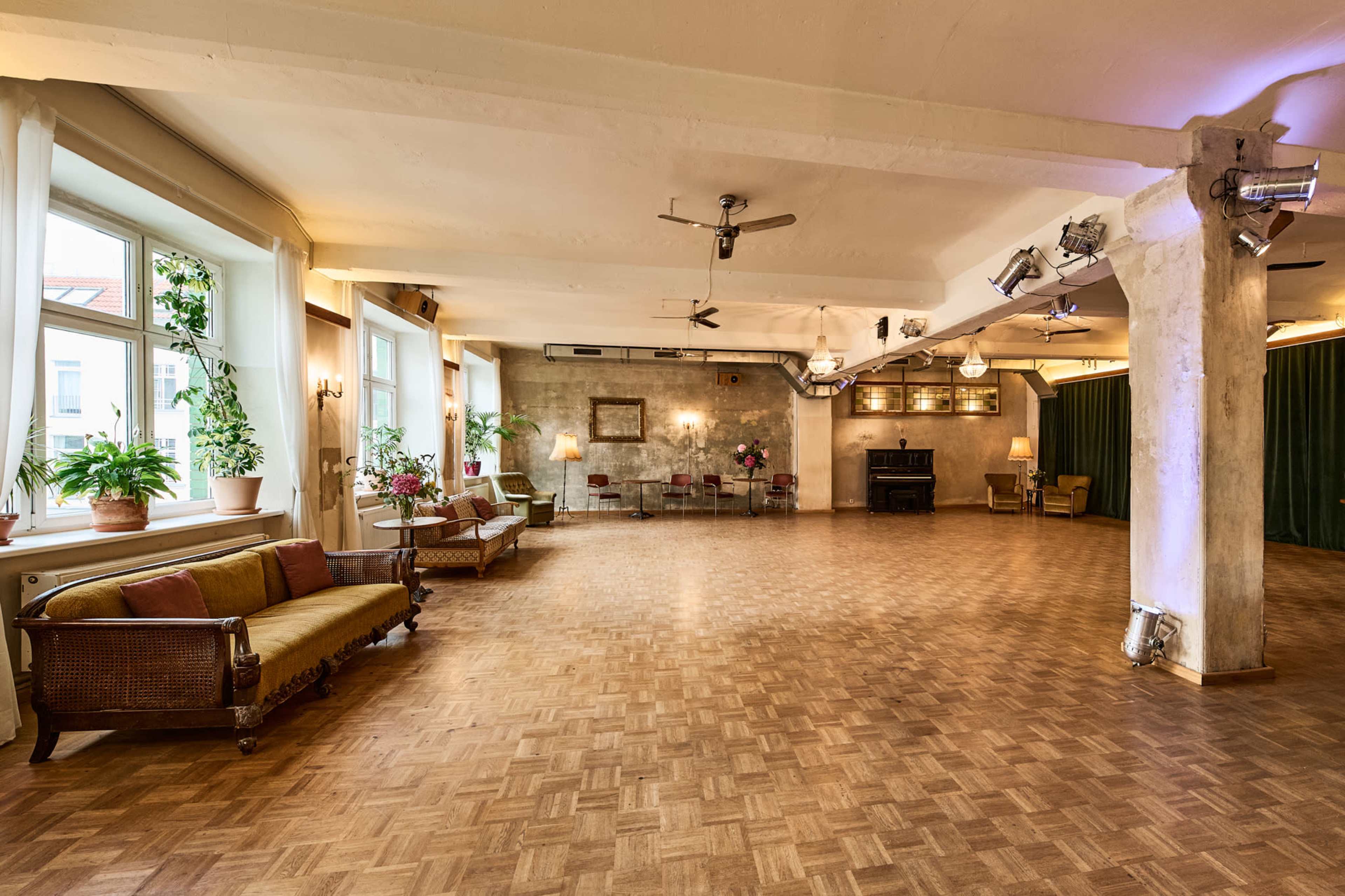 The image shows a spacious, empty room with wooden flooring, large windows, and decorative plants, featuring a vintage couch and a small stage area.