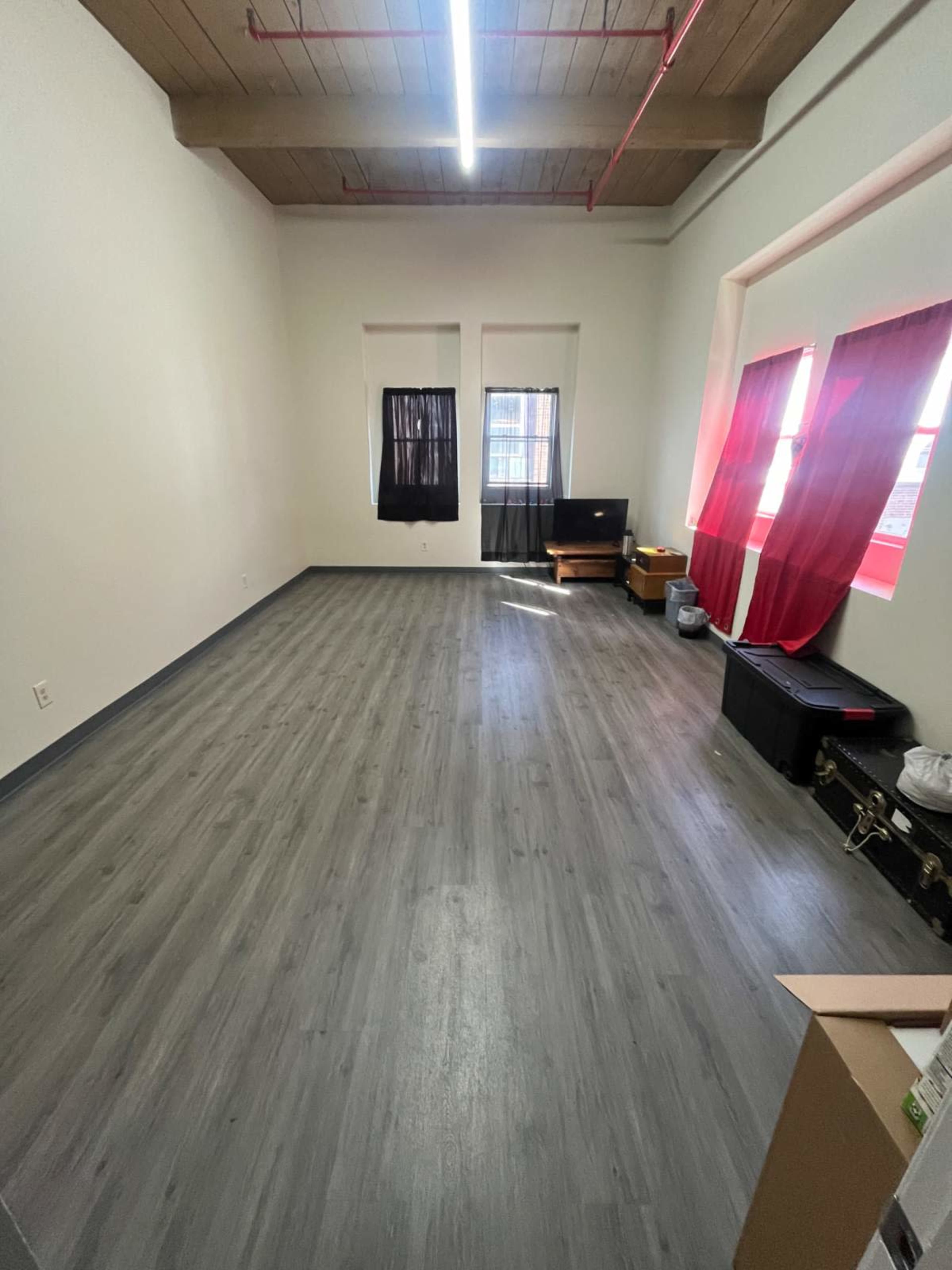 The image shows a spacious room with light gray flooring, two windows with black and red curtains, and minimal furniture including a television and a chest.