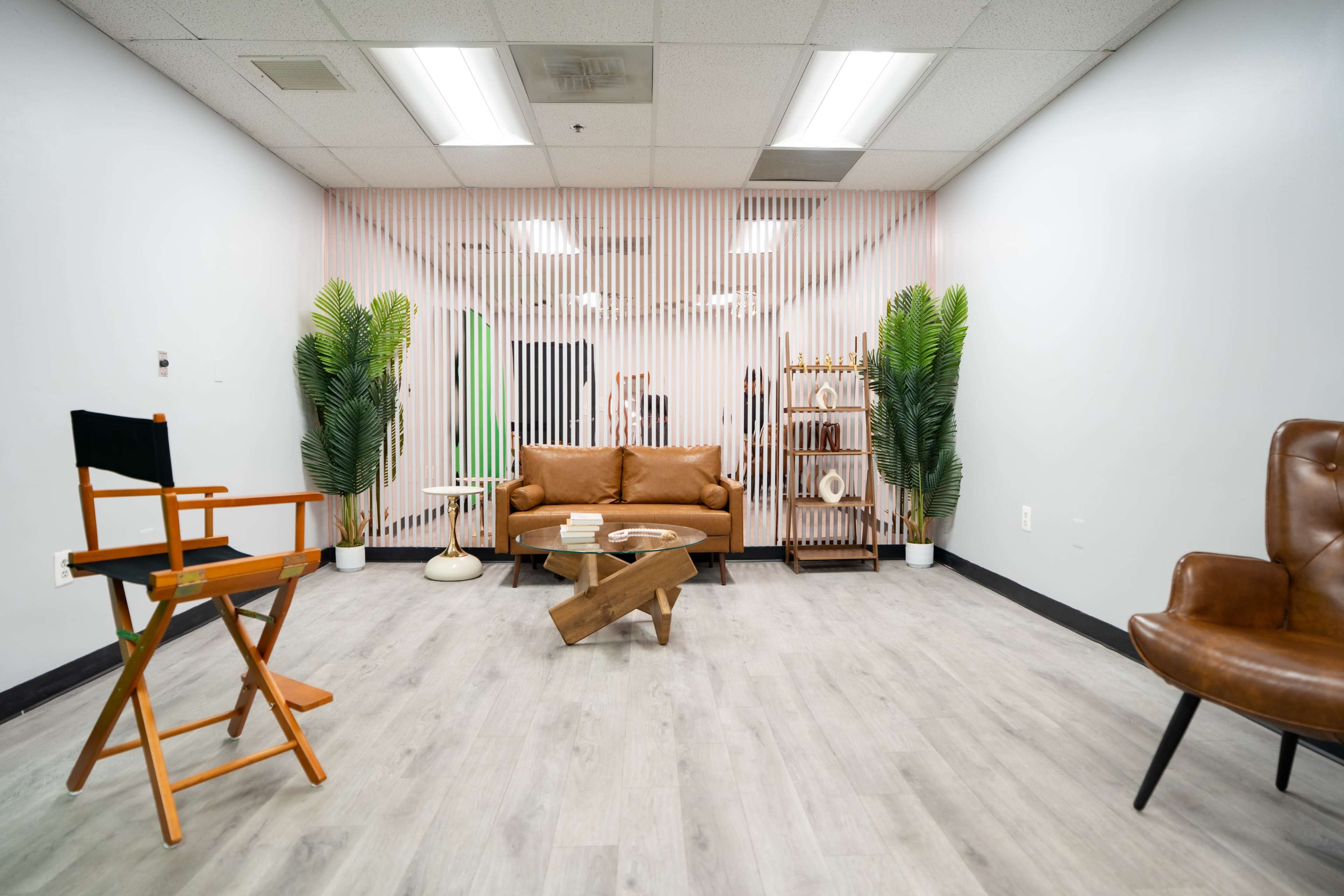 The image shows a modern interior space with a brown couch, a wooden coffee table, a director's chair, and a leather armchair, surrounded by potted plants and a slatted wall.