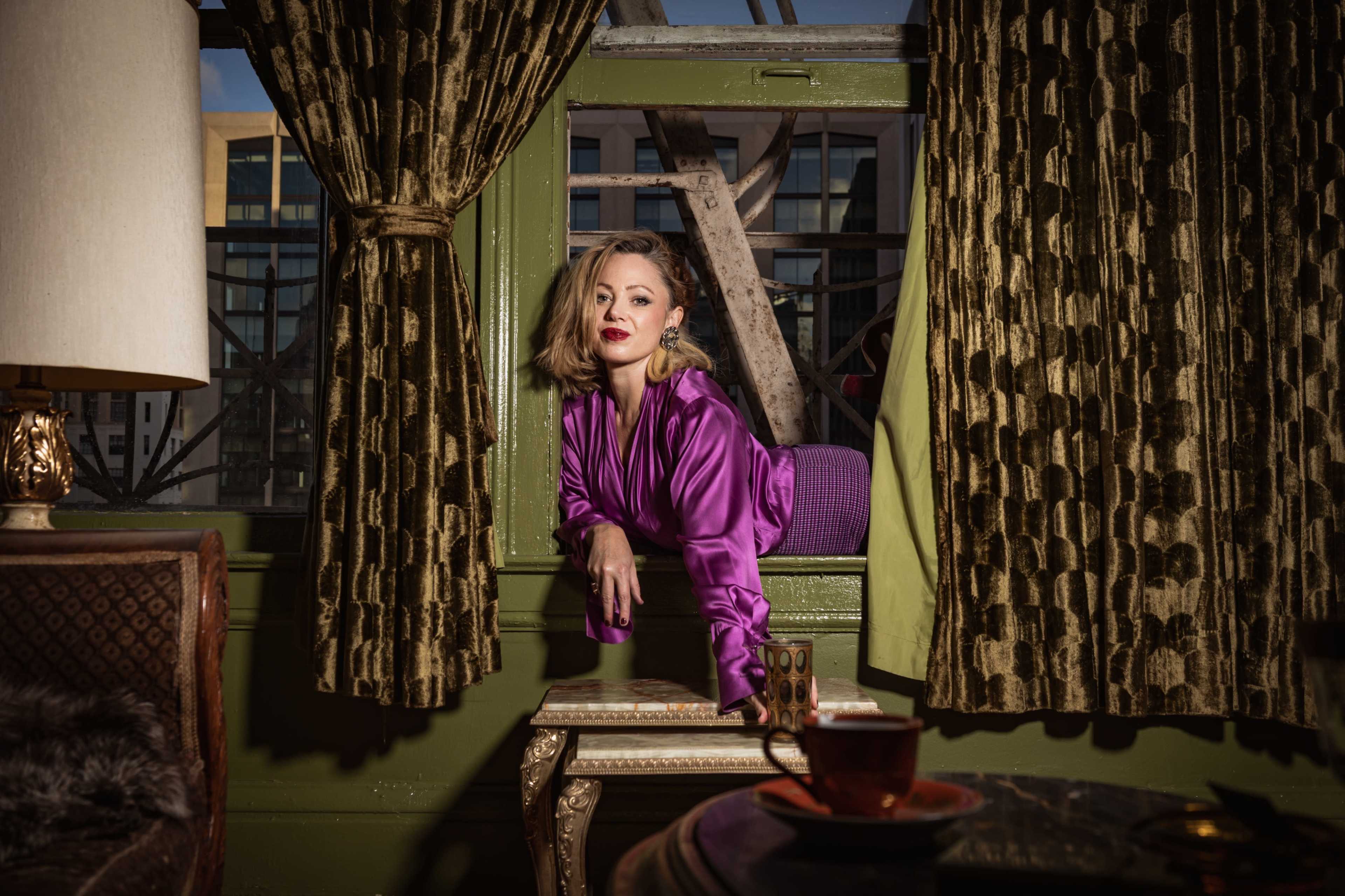 A woman in a purple satin robe leans out of a window surrounded by patterned curtains, with a vintage lamp and a small table in the foreground.
