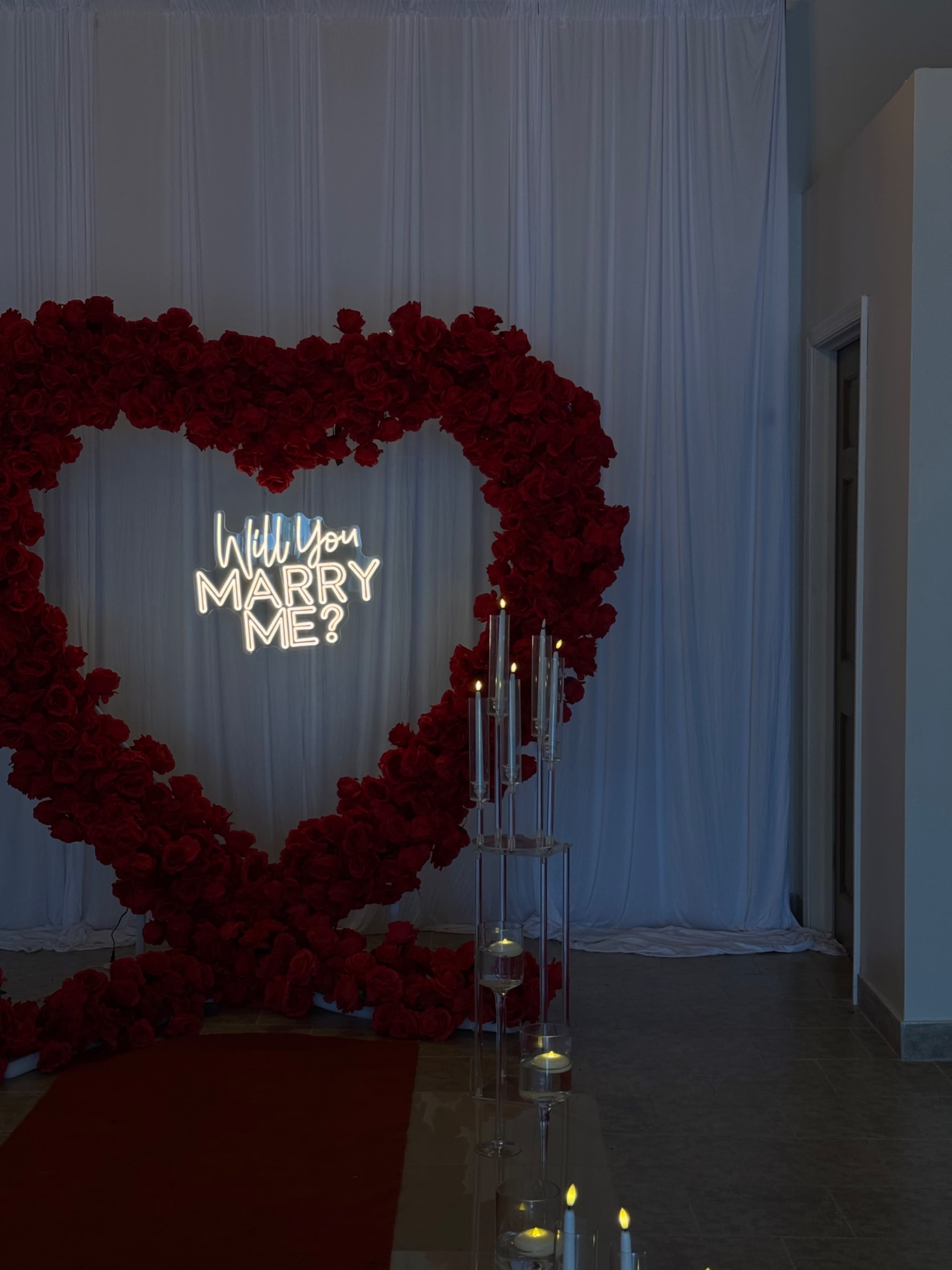 The image shows a romantic proposal setup featuring a large heart-shaped arrangement of red roses, a glowing sign that reads "Will You Marry Me?", and candles arranged on a clear display near a white backdrop.