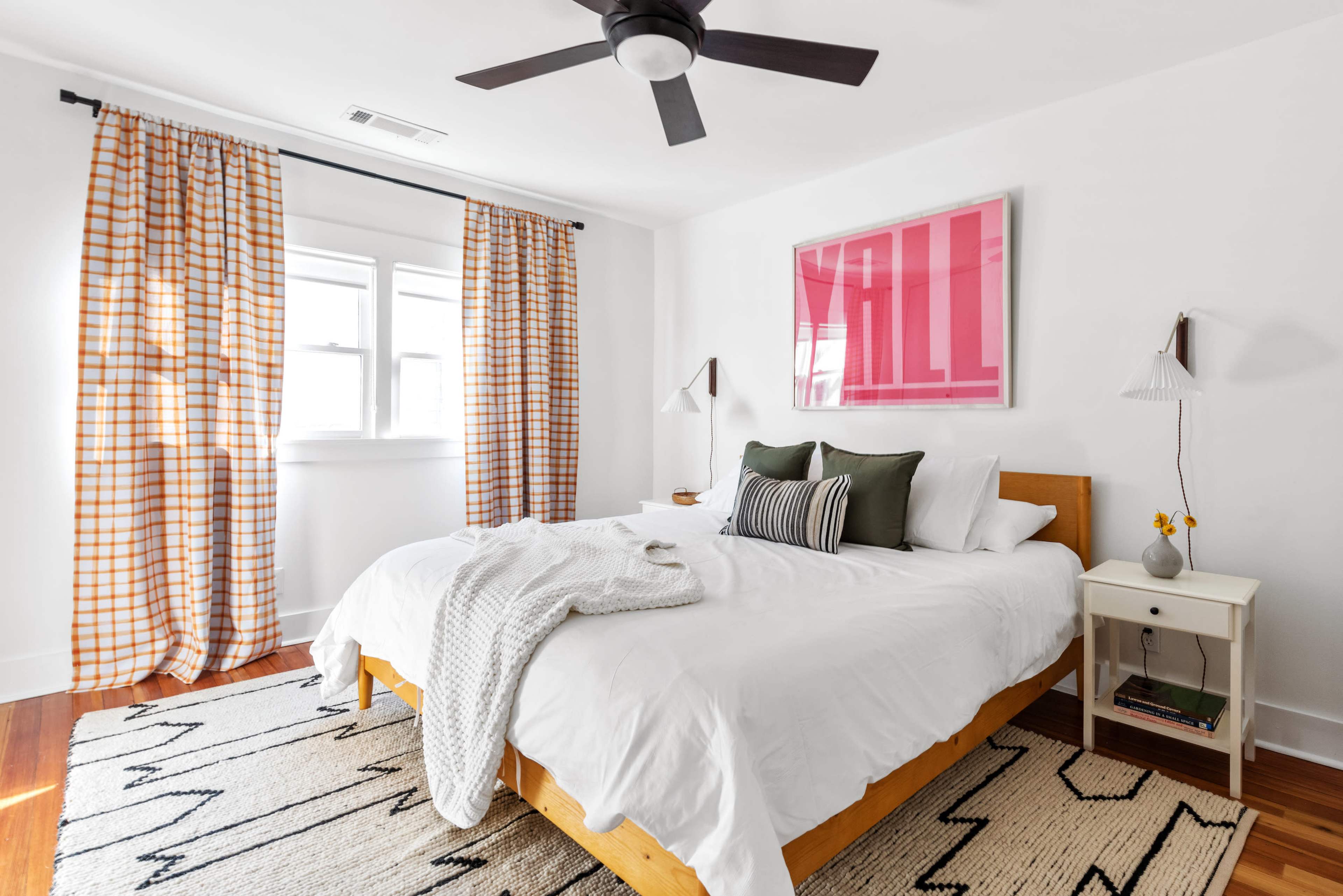 A bright bedroom features a large bed with white linens, a patterned throw blanket, and two bedside lamps, alongside windows dressed with checkered curtains.