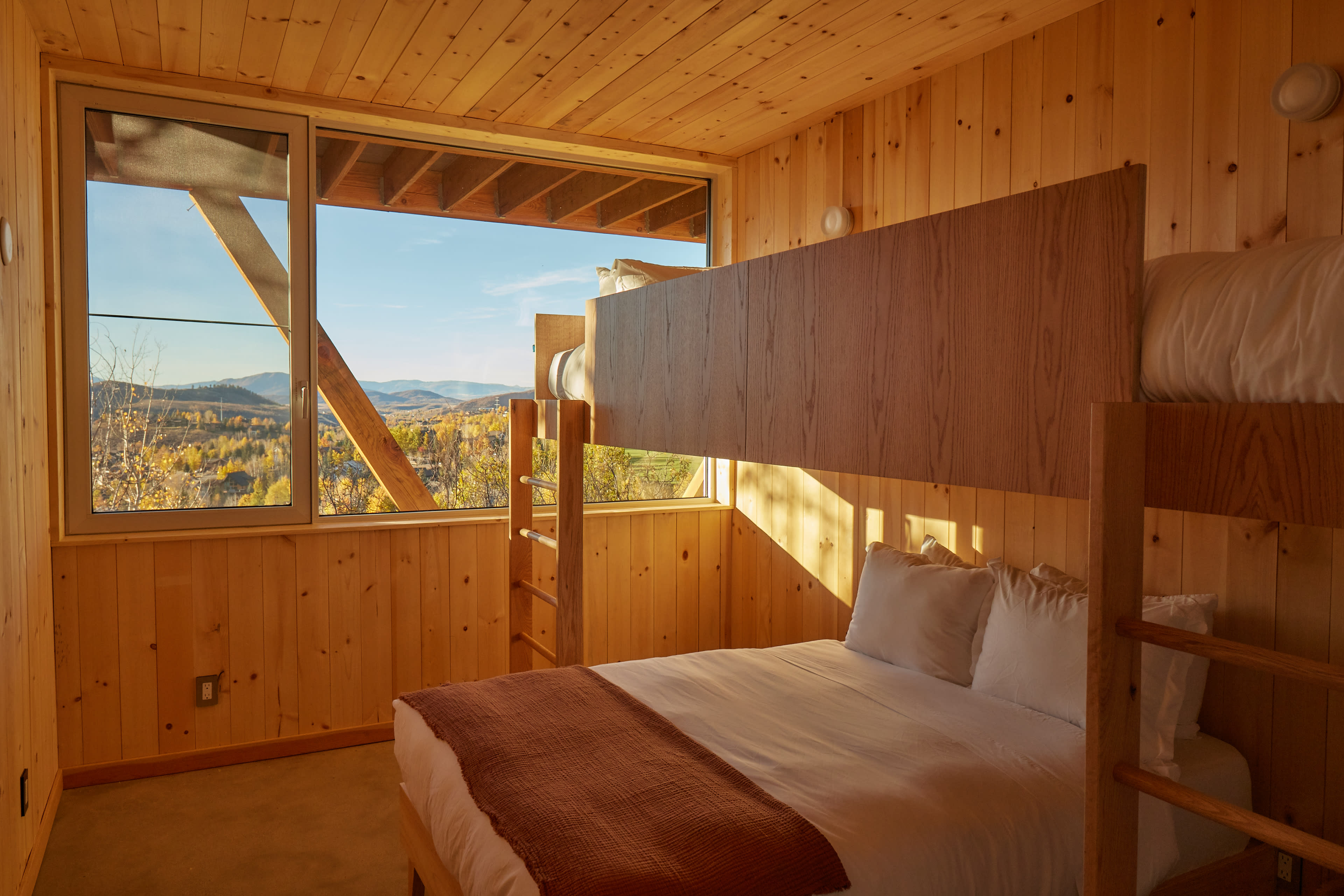 The image features a wooden-paneled bedroom with a large window showing a view of mountains, a double bed, and a bunk bed against the wall.