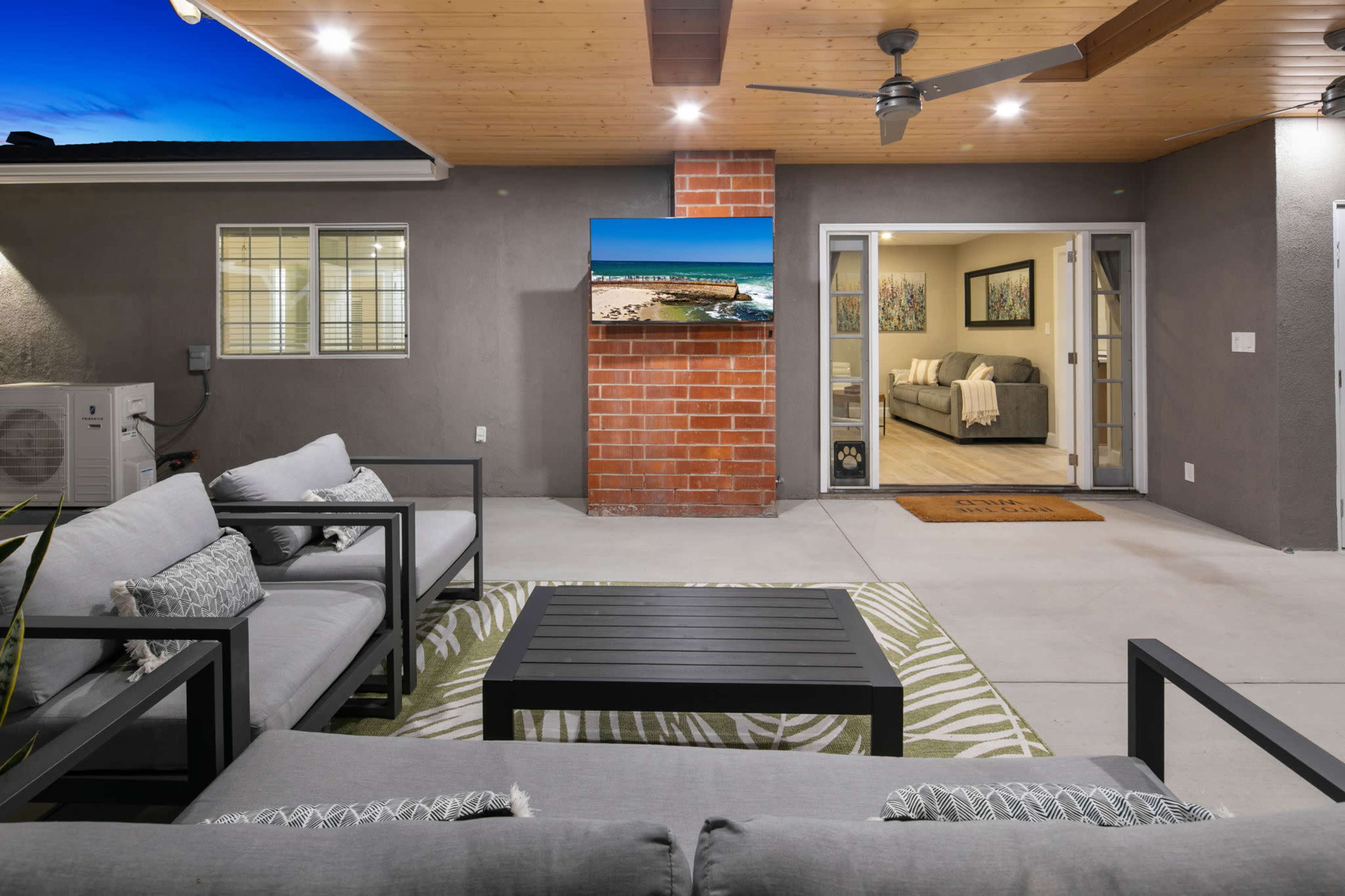 The image shows a modern outdoor living space with a seating arrangement, a coffee table, and a television mounted on a brick wall, adjacent to an interior room visible through large glass doors.
