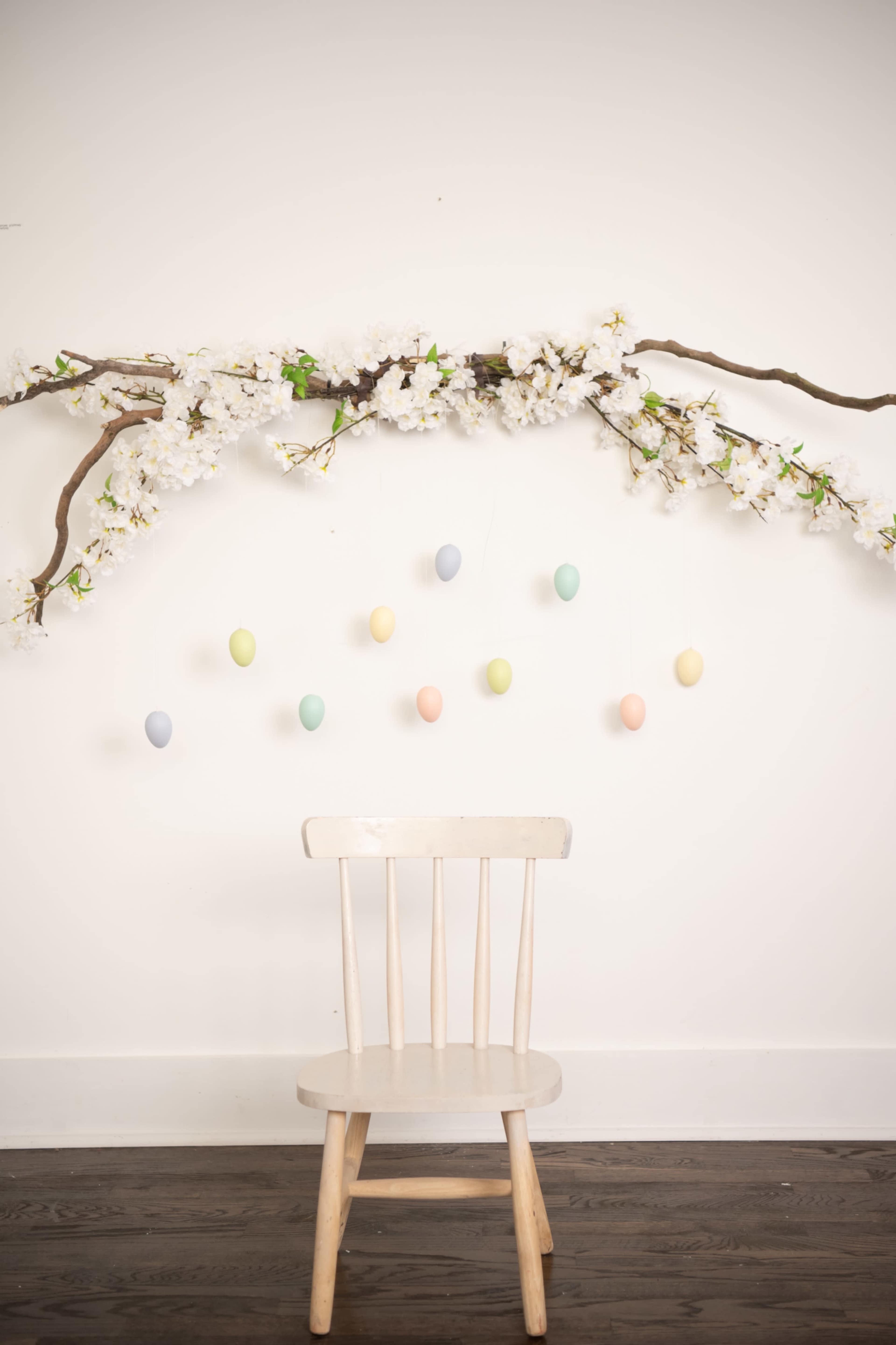 A simple wooden chair is placed underneath a decorative branch adorned with faux cherry blossoms and hanging pastel-colored eggs against a plain wall.