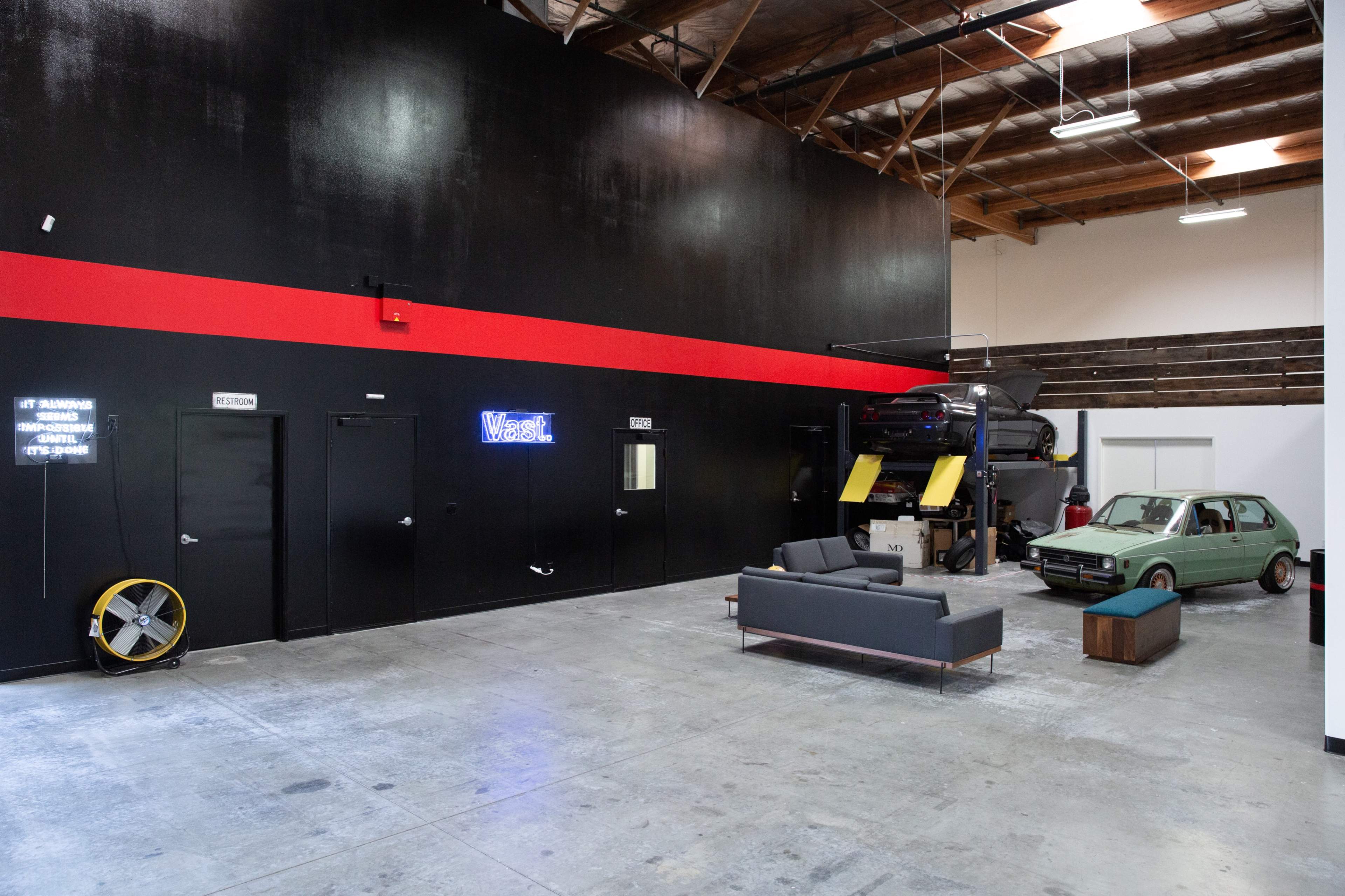 The interior of a garage features a gray couch and a green car, with black walls and a red stripe, alongside doorways and a neon sign.