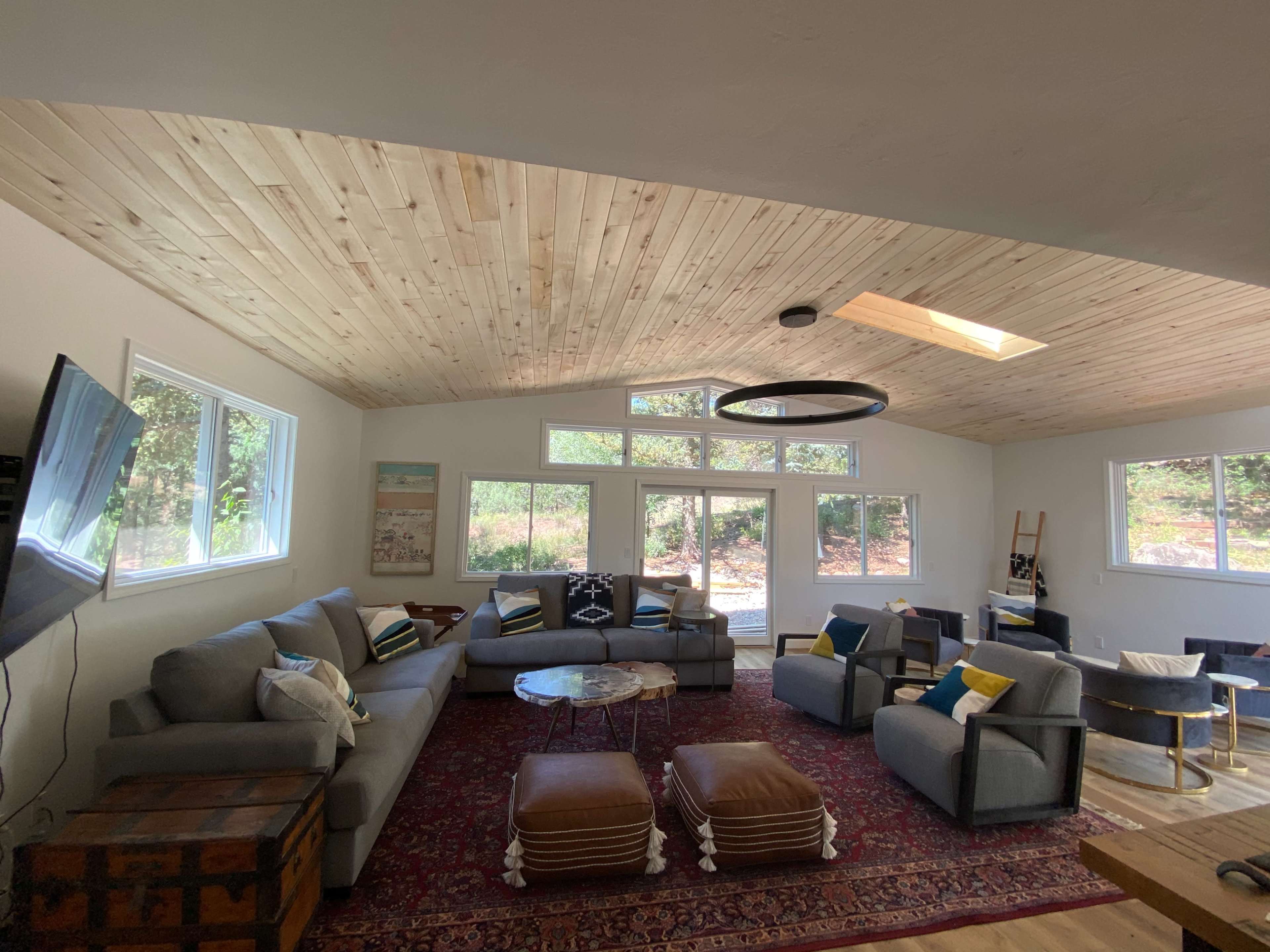 A spacious living room with a vaulted wooden ceiling, large windows, and a combination of couches and armchairs arranged around a central coffee table.