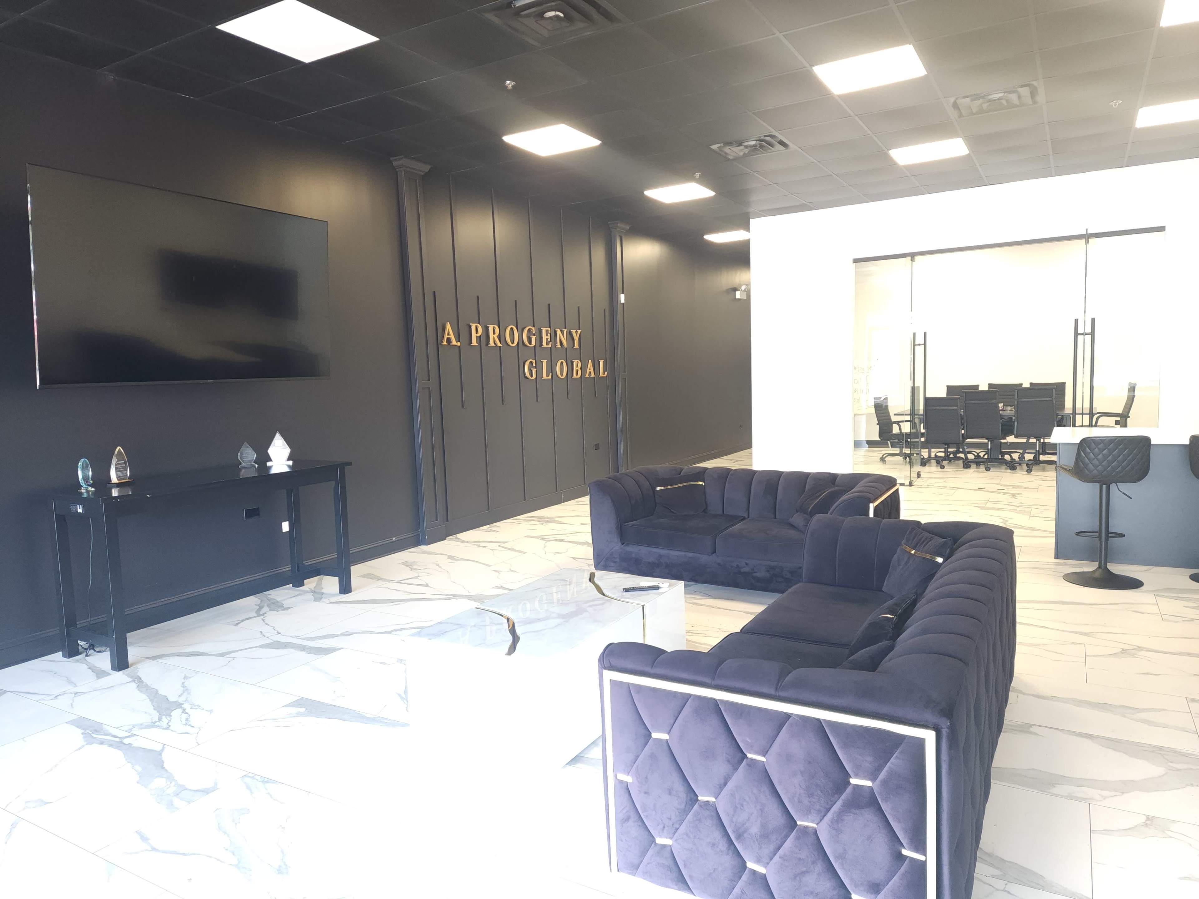 The image shows a modern office interior with a reception area featuring dark walls, two plush sofas, a coffee table, and a large screen on one wall, while a conference room is visible in the background.
