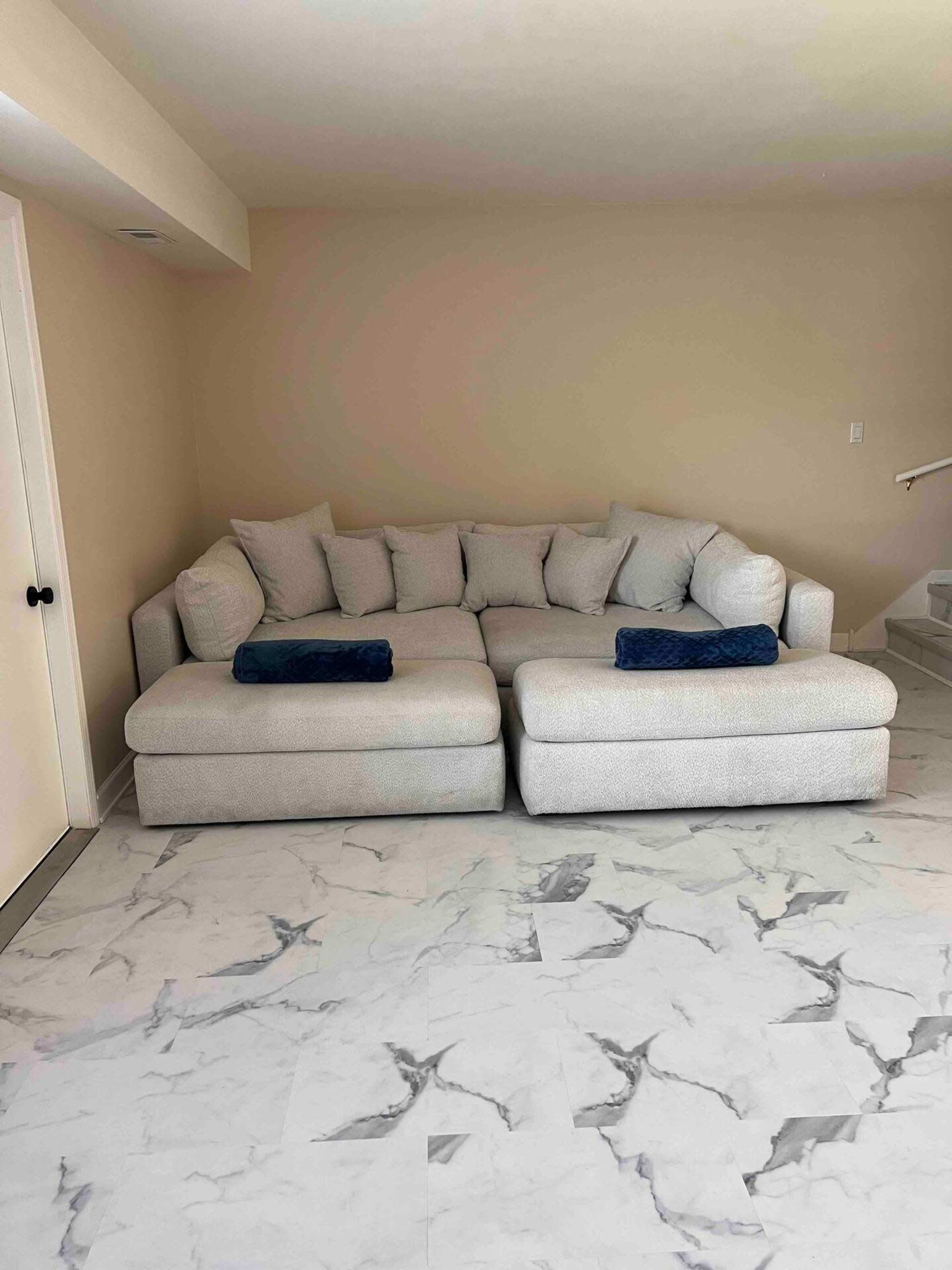A light-colored sectional sofa with several cushions is positioned on a marbled floor in a room with beige walls.