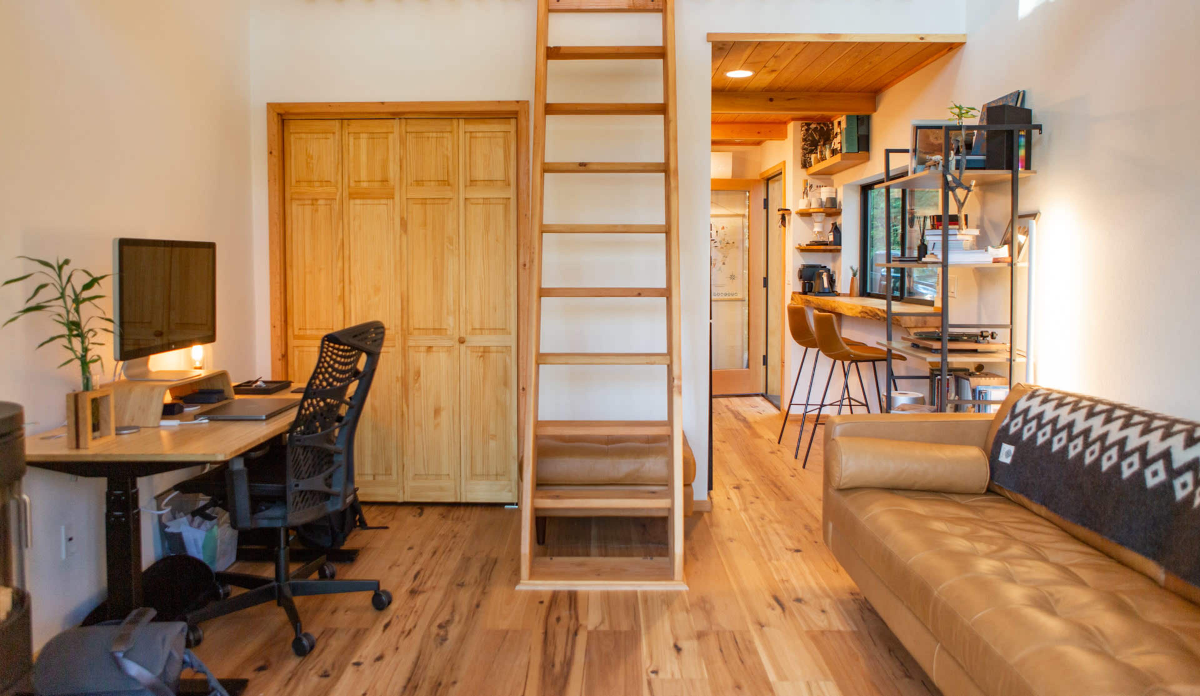 A small living space featuring a wooden ladder leading to an upper area, a desk with a computer, a sofa, and a kitchen area with stools.