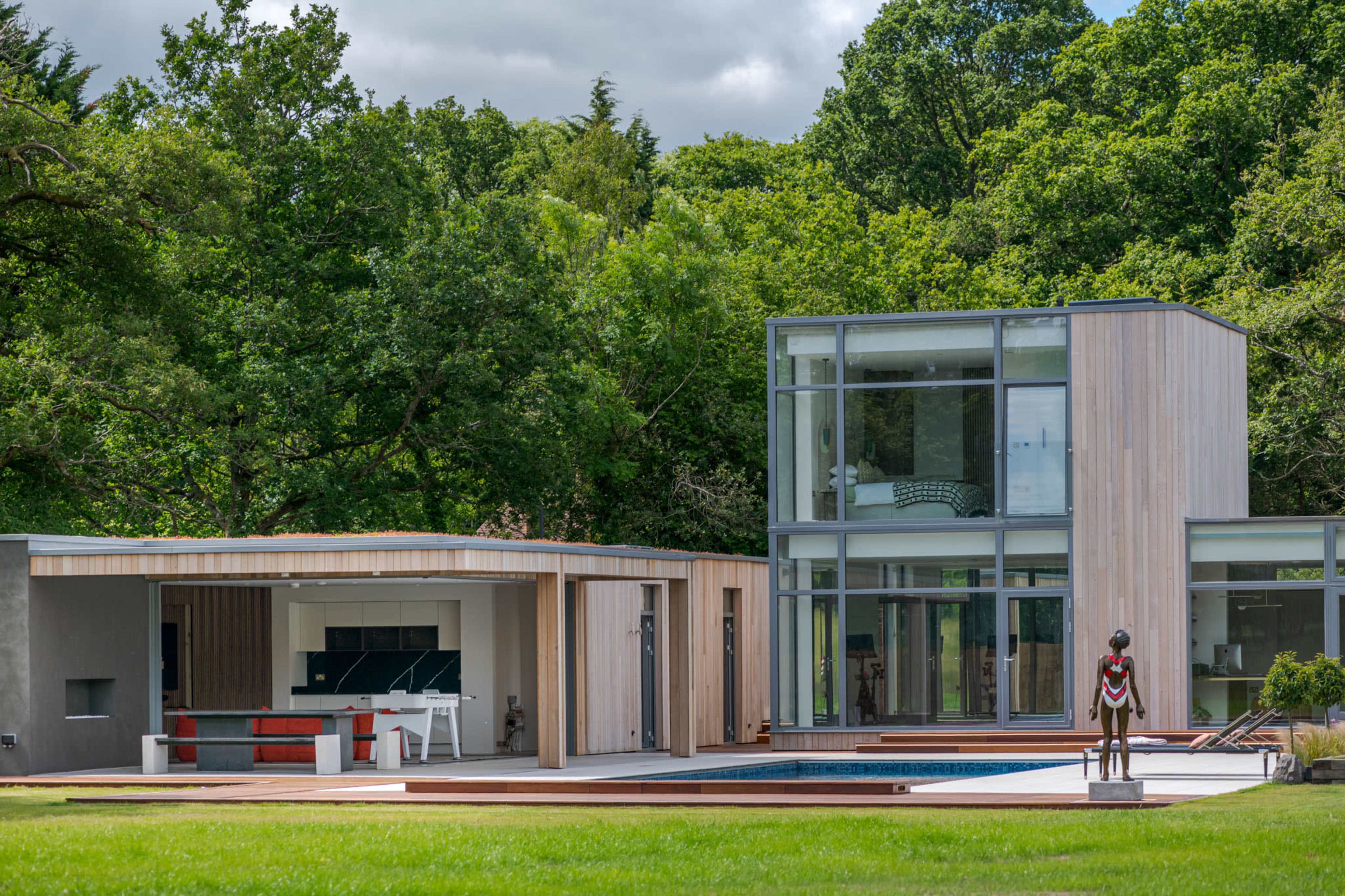 The image shows a modern house with large glass windows, a wooden exterior, and a swimming pool surrounded by a grassy area and lush trees.