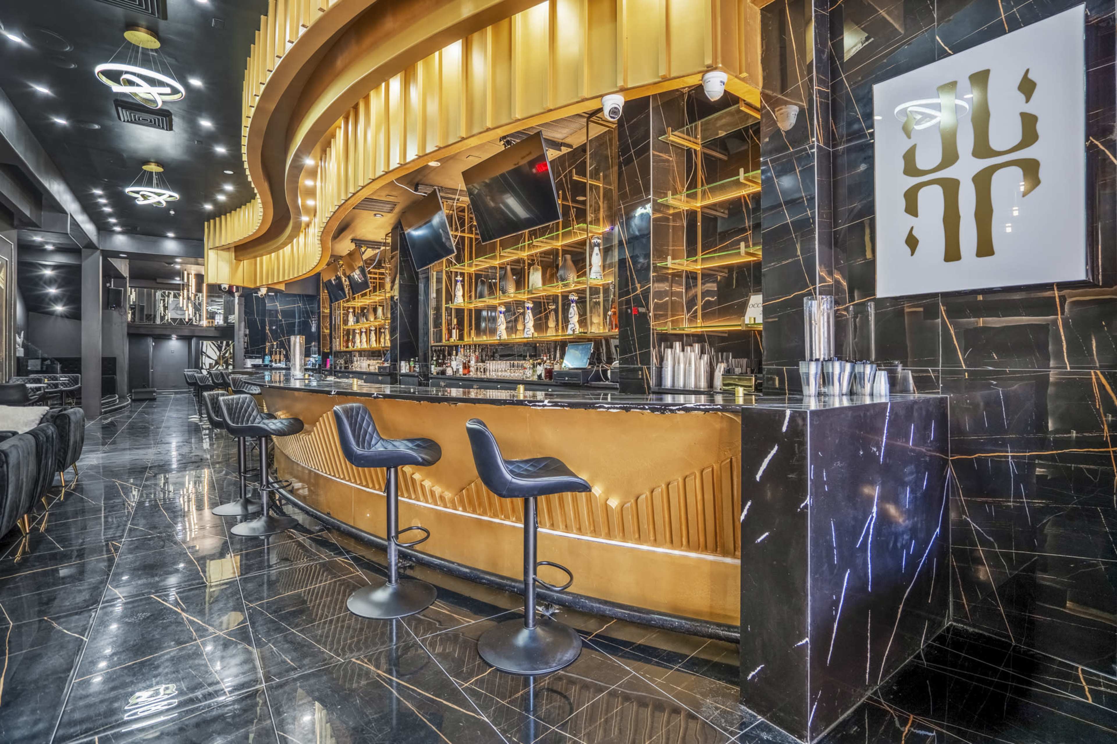A luxurious bar with a golden counter and black marble flooring, featuring several bar stools and shelves stocked with various bottles.