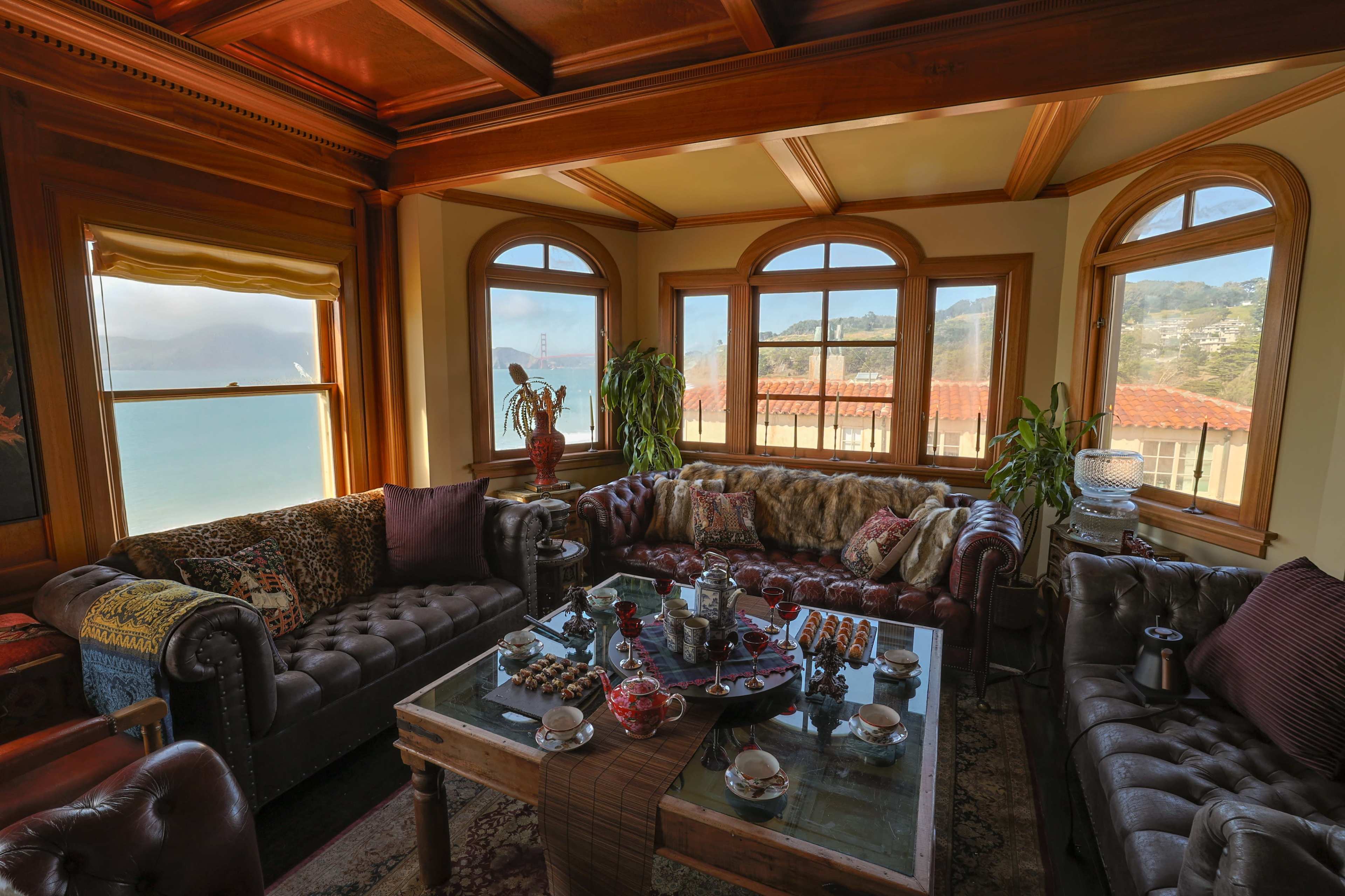 The image shows a cozy living room with large windows, a sectional couch, and a coffee table adorned with various items, overlooking a scenic view of the ocean and hills.