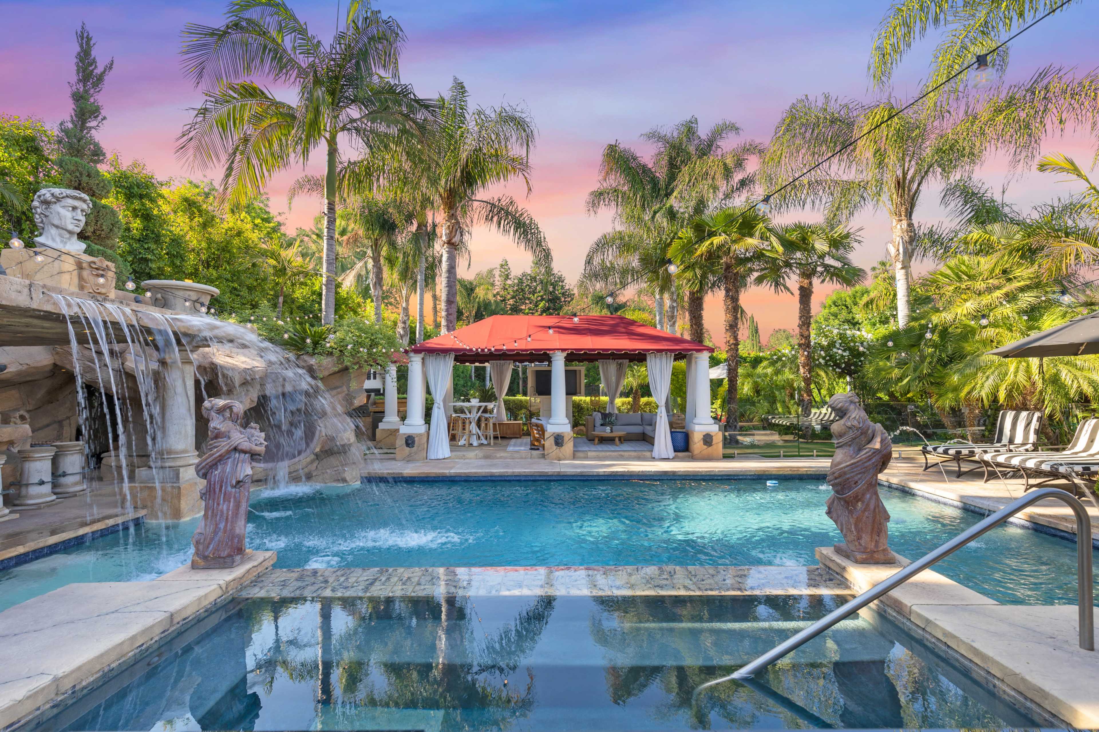 A luxurious outdoor pool area with a waterfall, palm trees, and a gazebo surrounded by patio furniture.