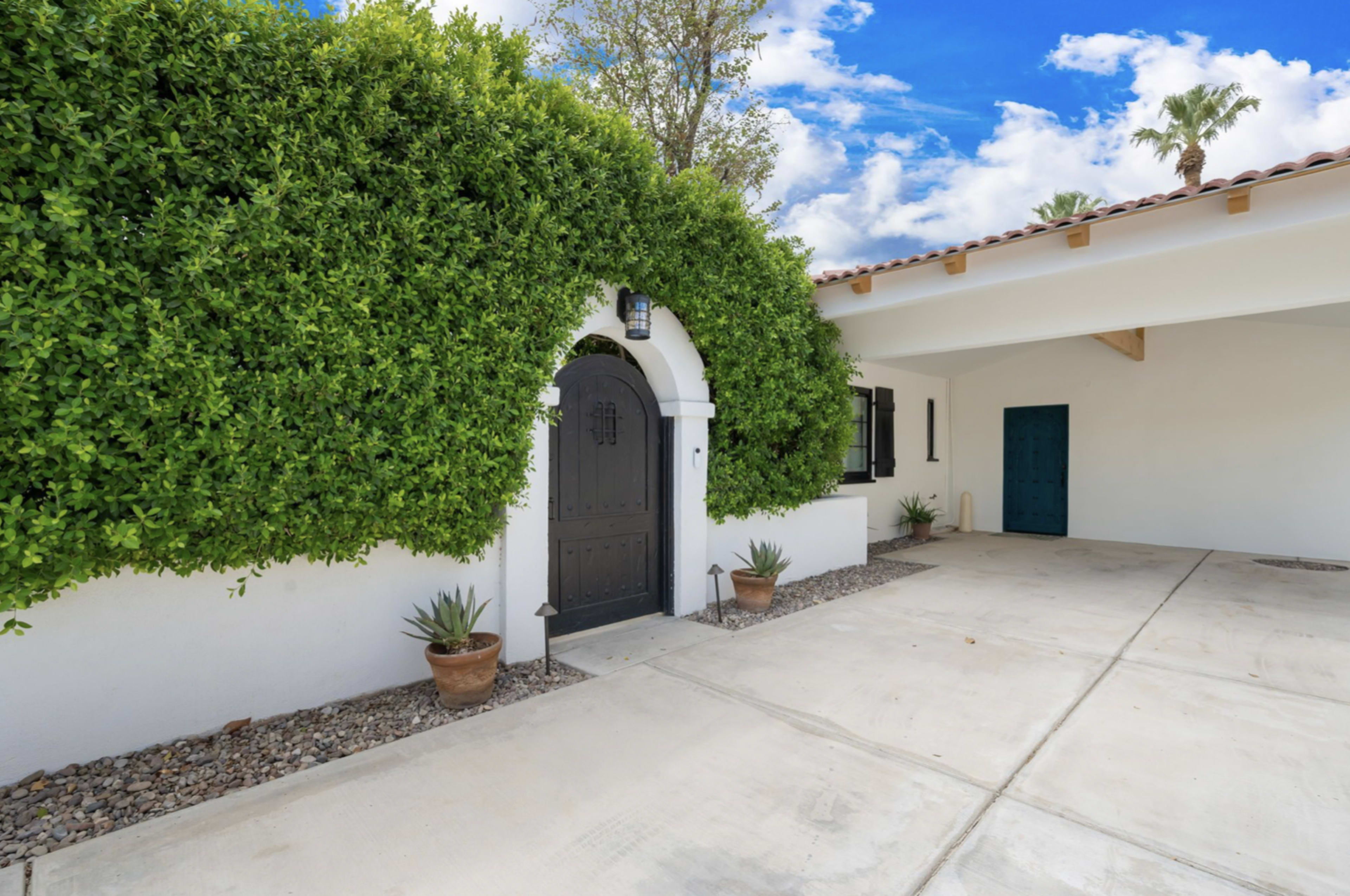 A white house with a dark wooden door framed by a lush green hedge and two potted plants on a concrete driveway.