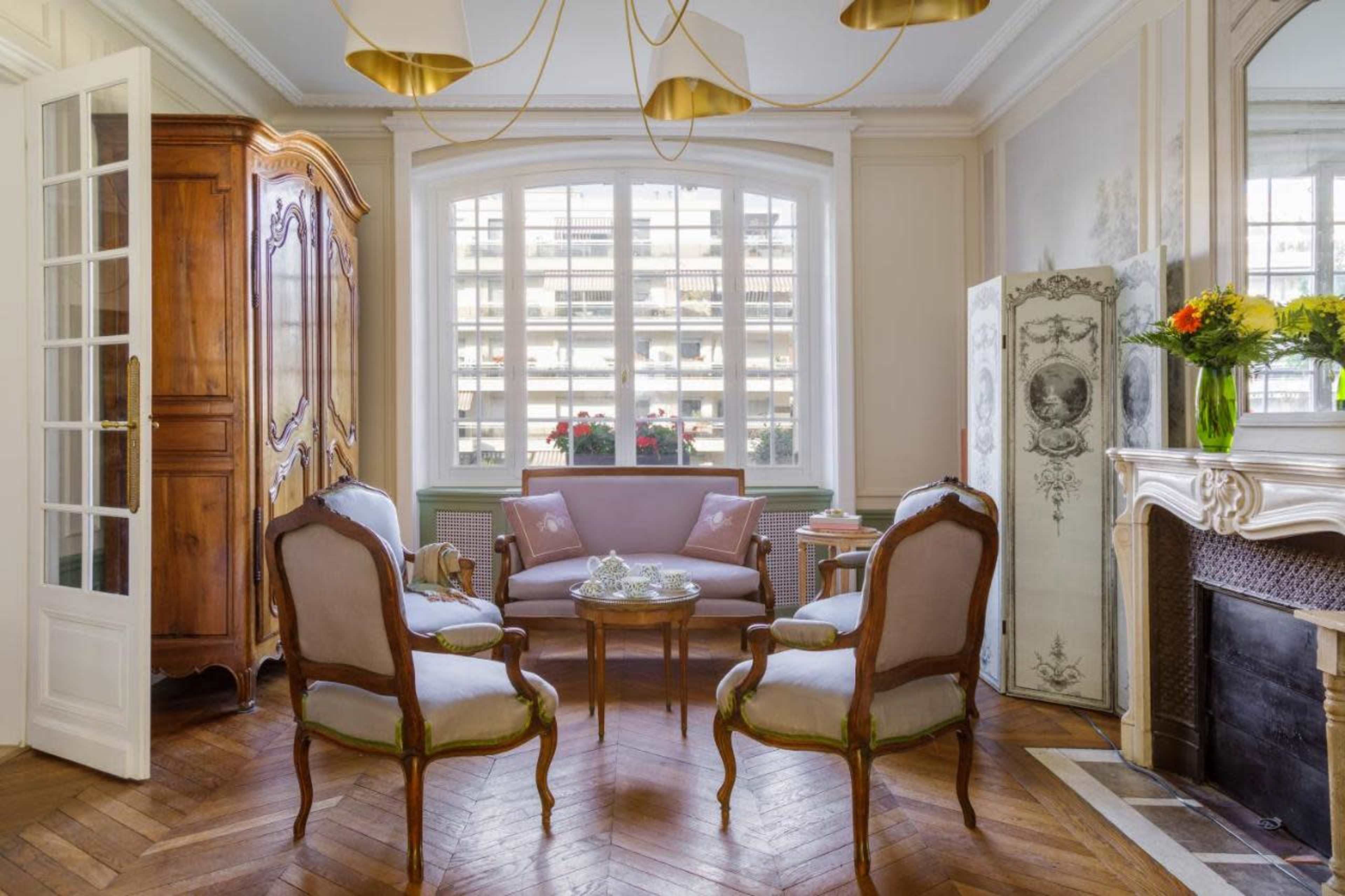A tastefully decorated living room featuring a wooden armoire, a round table with glassware, and elegant chairs arranged around it, all set in front of a large window.