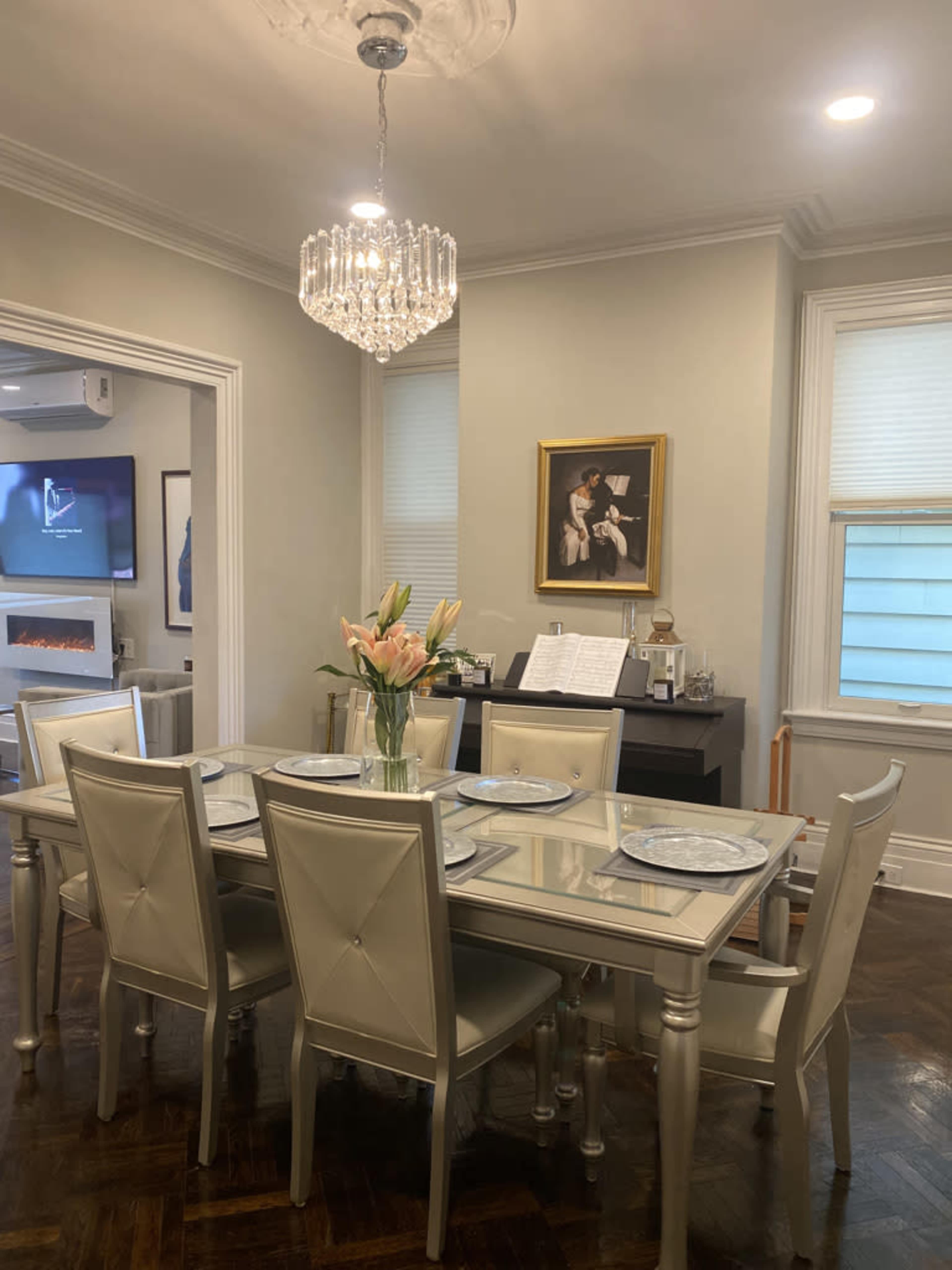 A dining room features a glass-top table surrounded by six upholstered chairs, with a chandelier above and a painting on the wall.