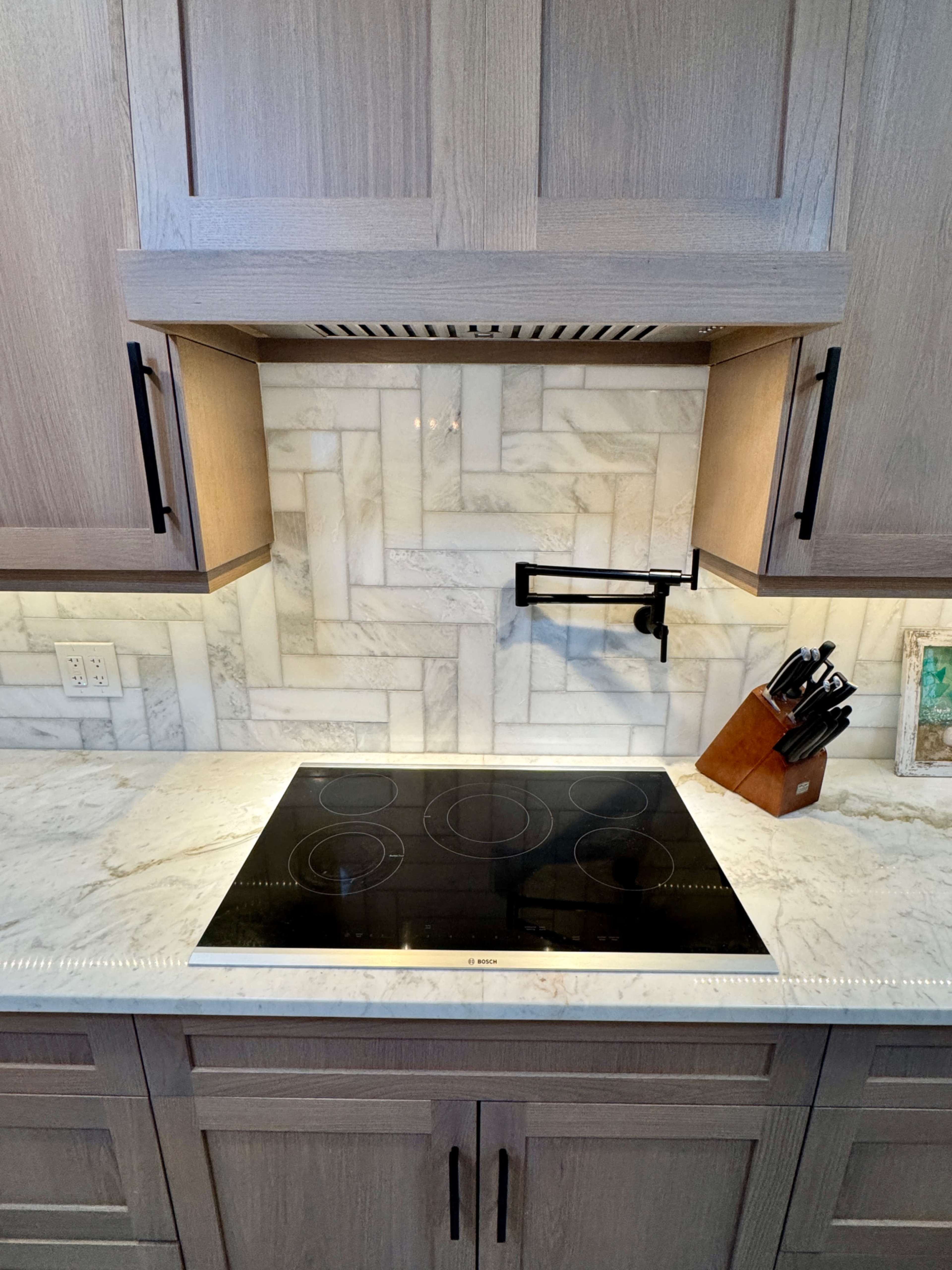 A modern kitchen features a sleek black induction cooktop beneath a wooden hood with a textured marble backsplash.