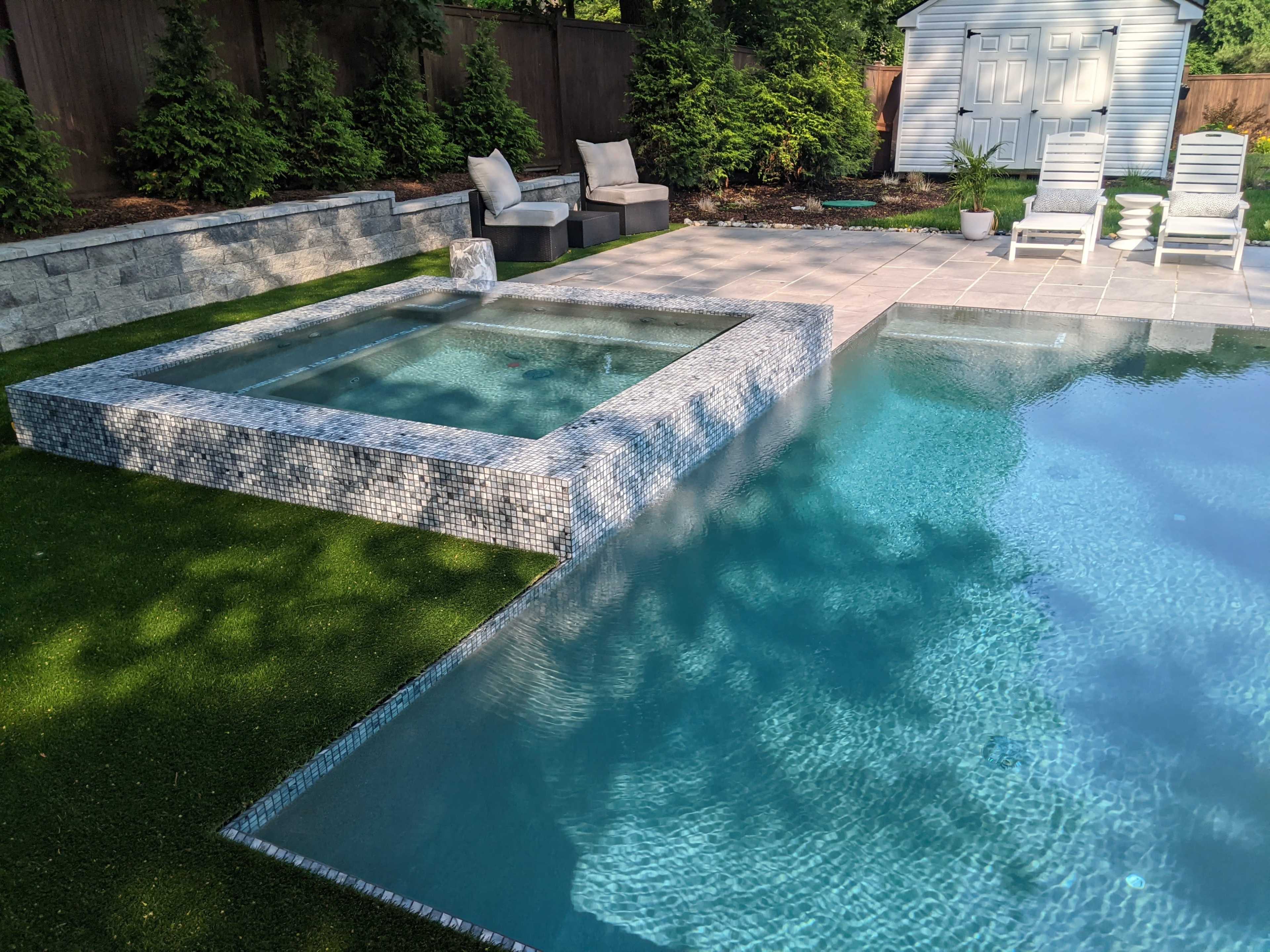 A modern backyard featuring a swimming pool with an attached spa, surrounded by stone pavers and artificial grass.