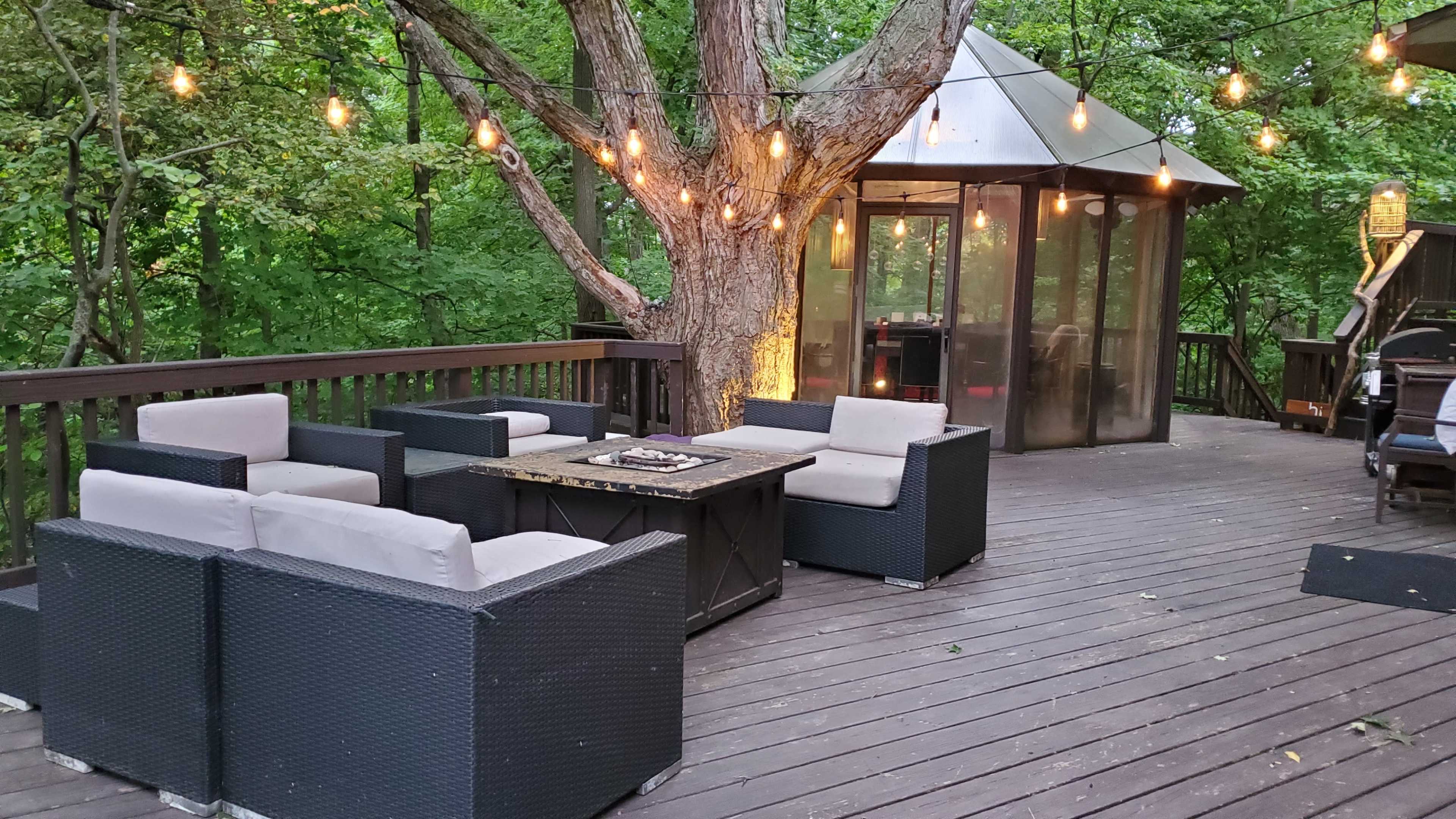 A wooden deck with four seating chairs arranged around a central table, decorated with string lights and surrounded by trees.