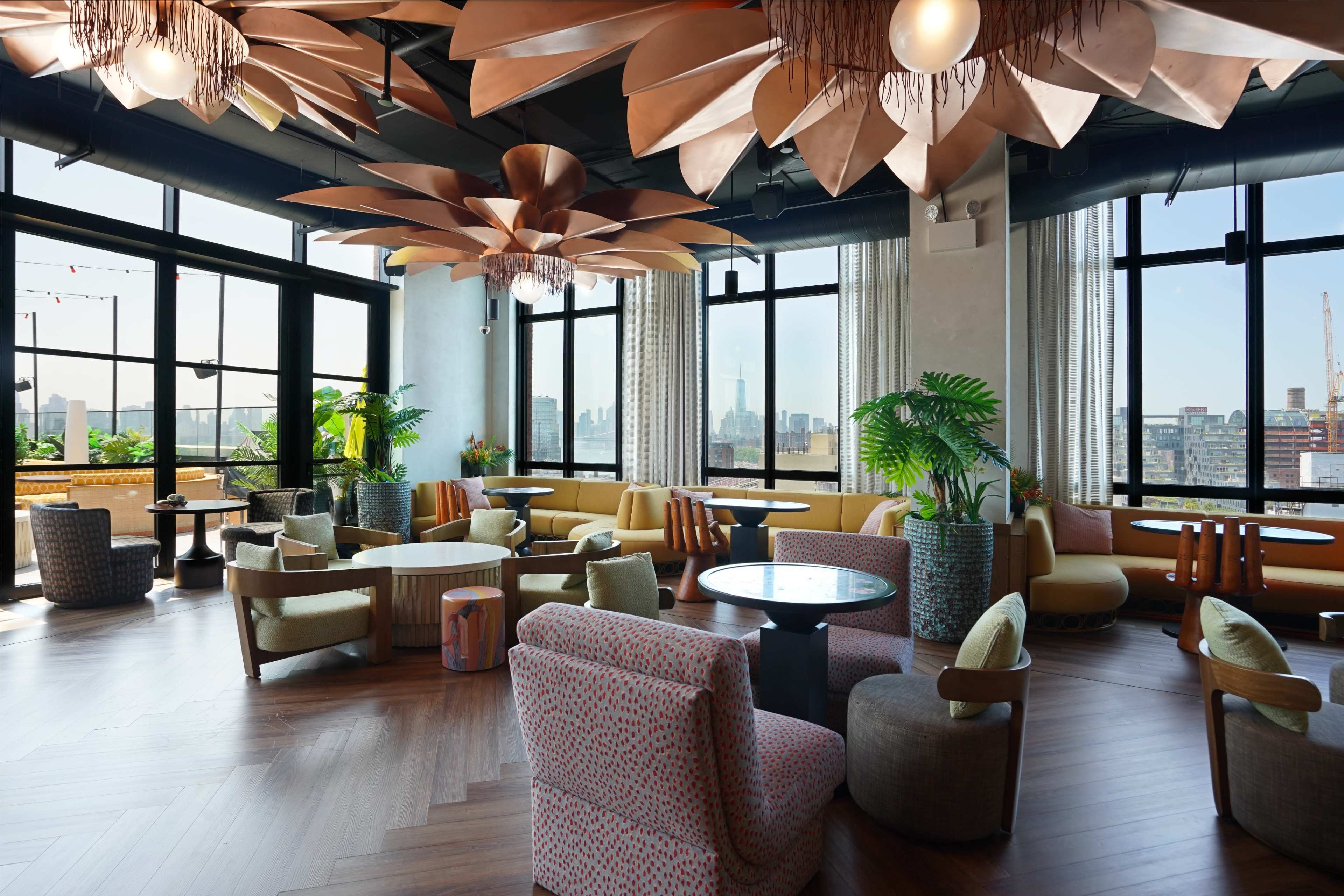 The image shows a modern lounge area with stylish seating, large windows offering a city view, and decorative floral light fixtures.