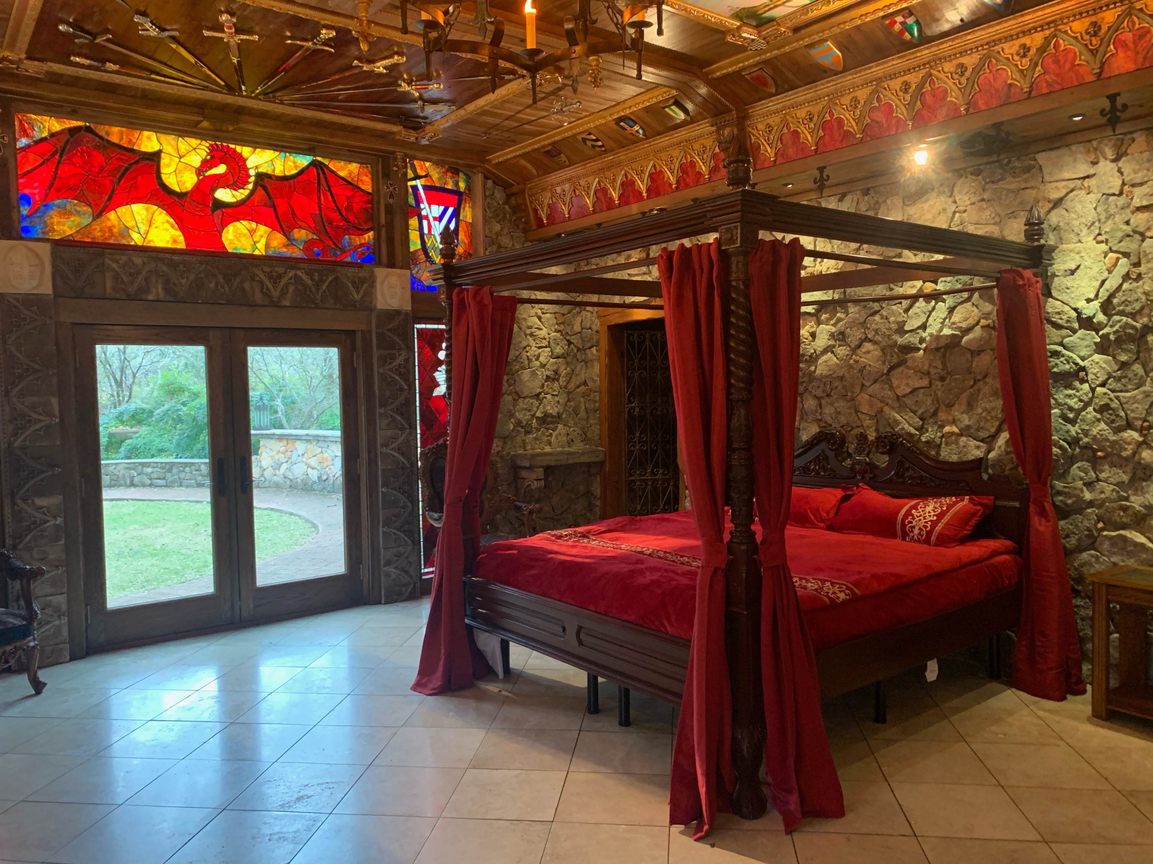 A four-poster bed with red drapes in a stone-walled bedroom featuring stained glass windows and a glass door leading to an outdoor area.