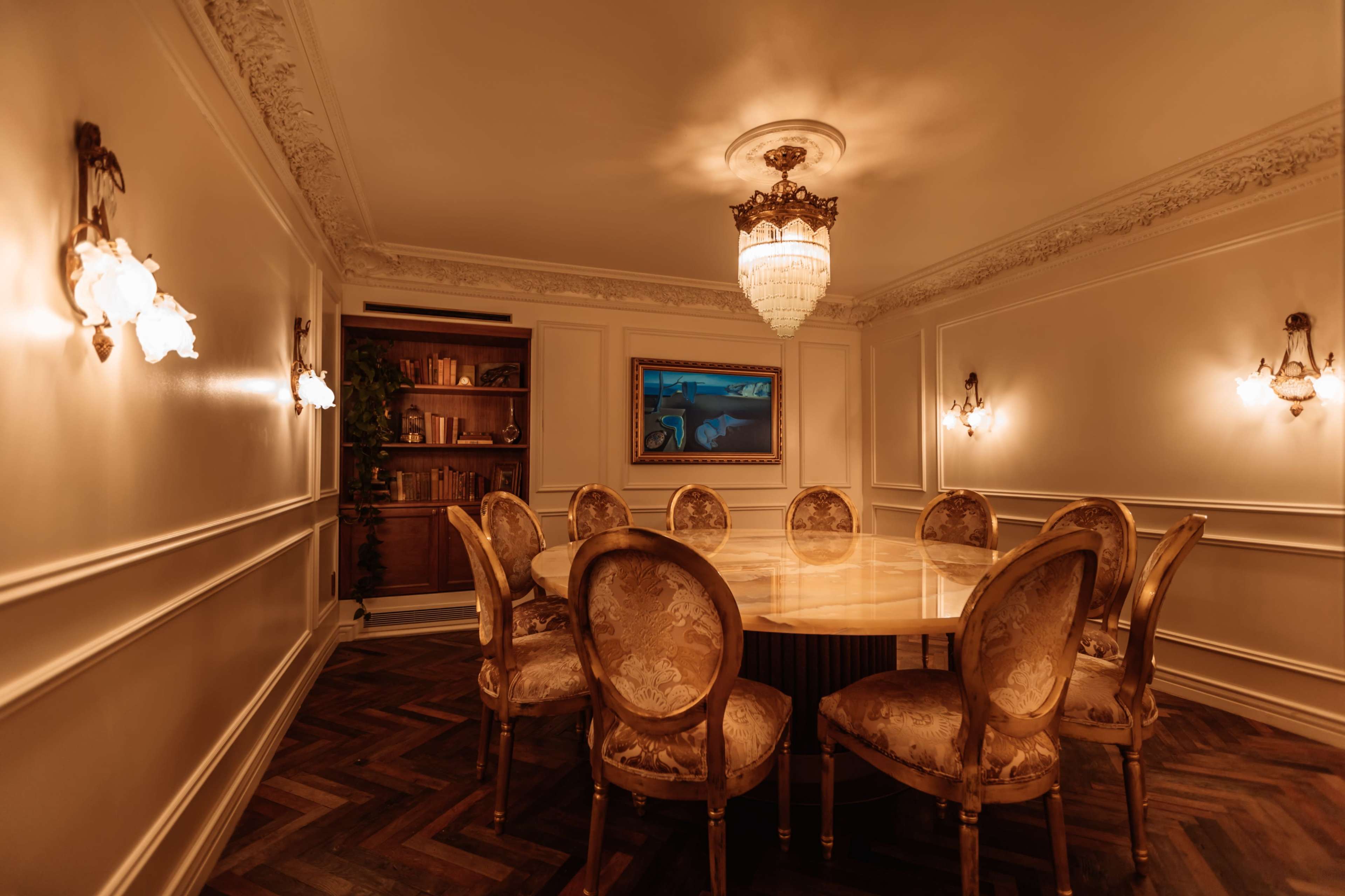 The image shows an elegantly designed dining room with a round table surrounded by ornate chairs, illuminated by a chandelier and wall sconces.