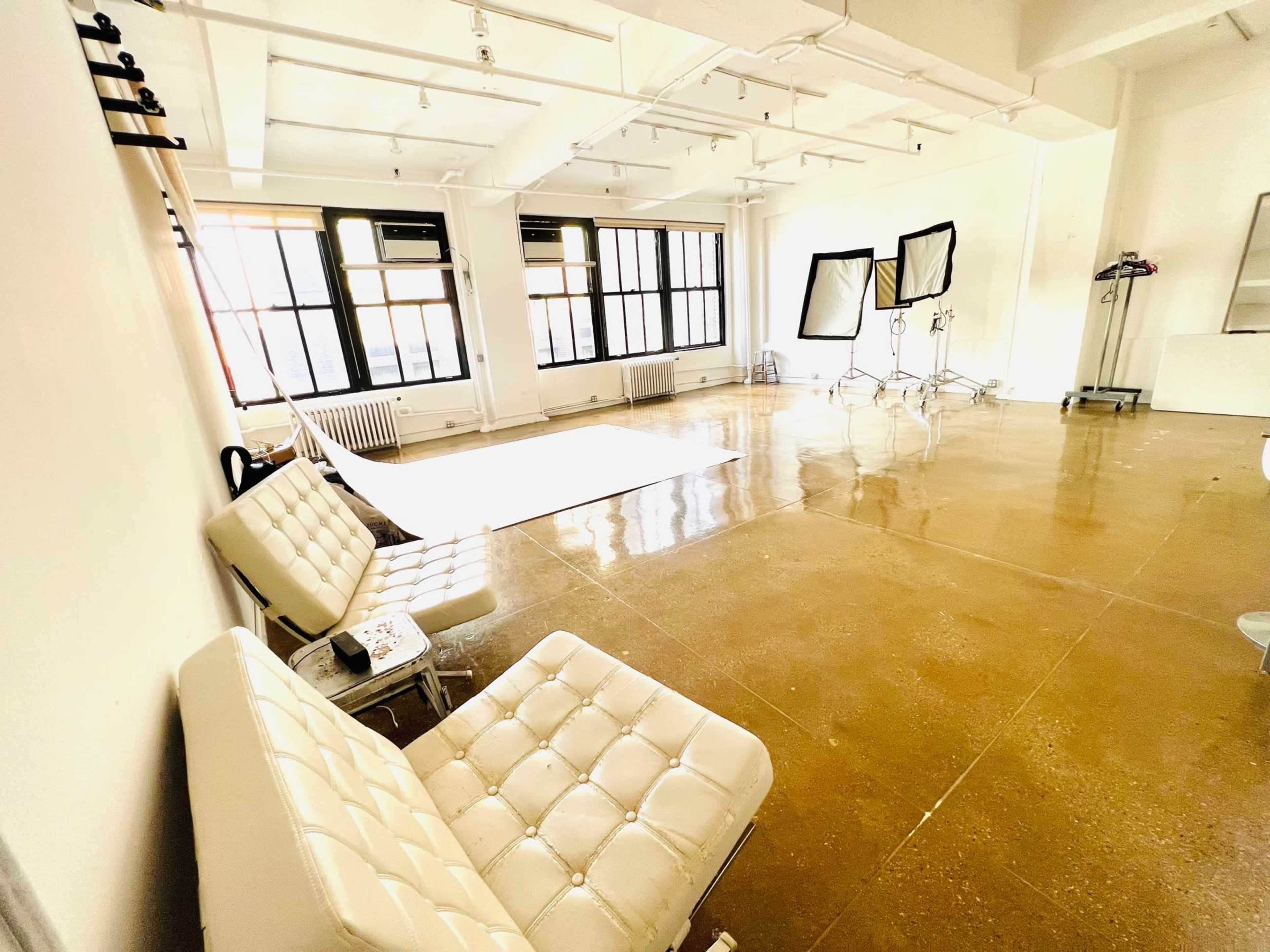The image shows a spacious, empty studio with polished concrete floors, large windows, and minimal furnishings, including two white chairs and several lighting equipment stands.