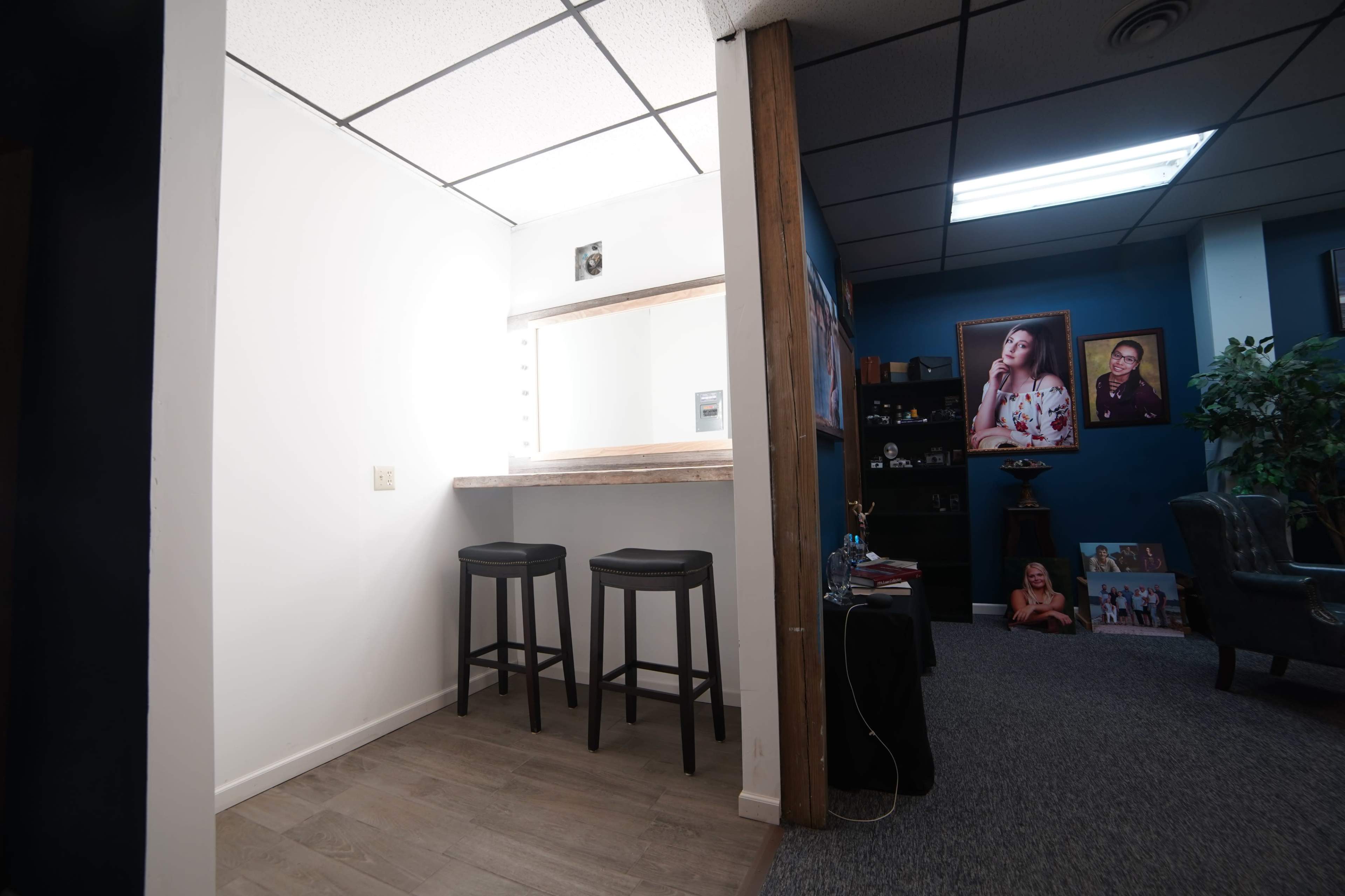 The image shows a small waiting area with two black stools and a mirror, adjacent to a room featuring a portrait and additional seating.