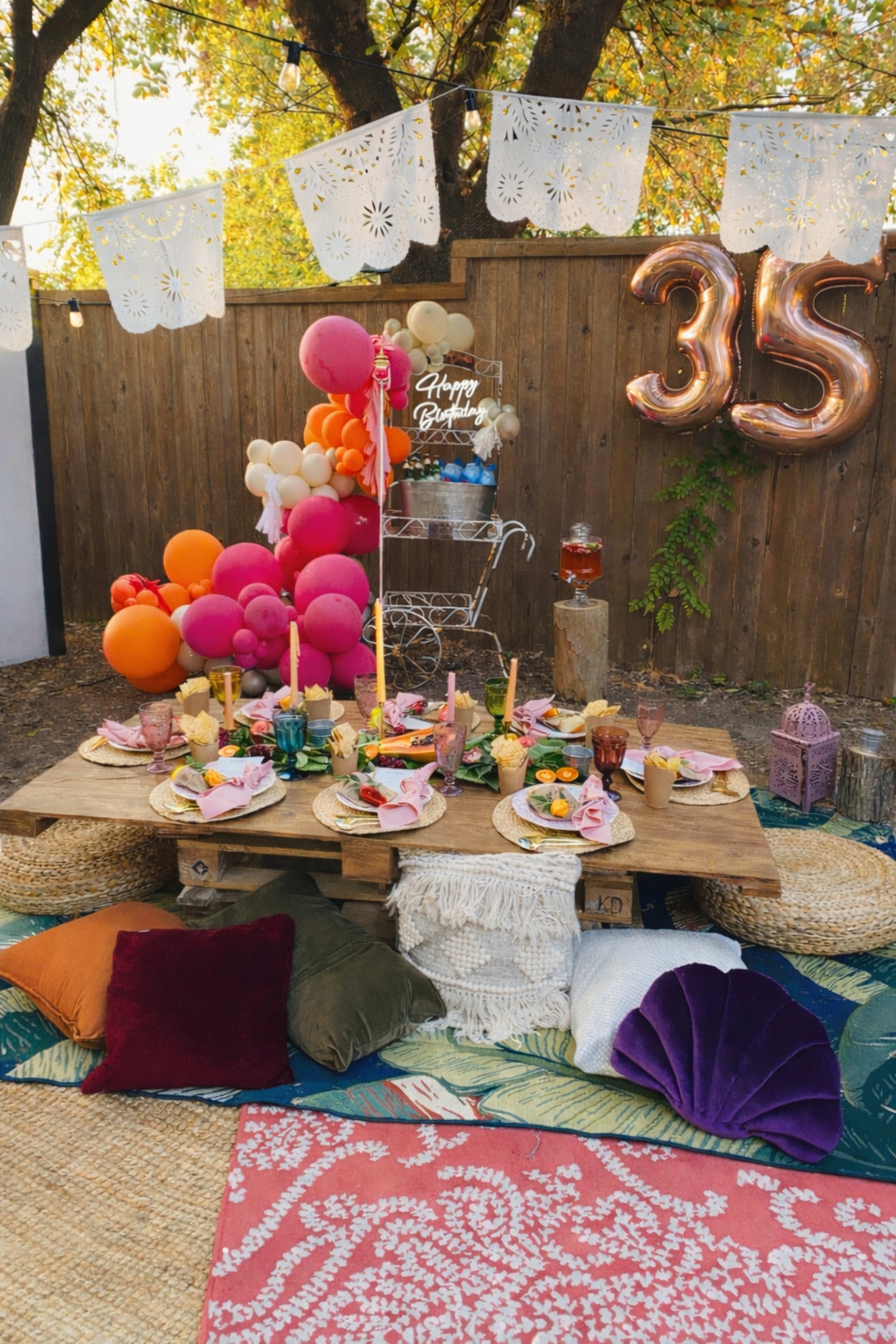 A rustic outdoor dining setup features a low wooden table adorned with colorful plates, candles, and decorations, surrounded by throw pillows and balloons for a birthday celebration.