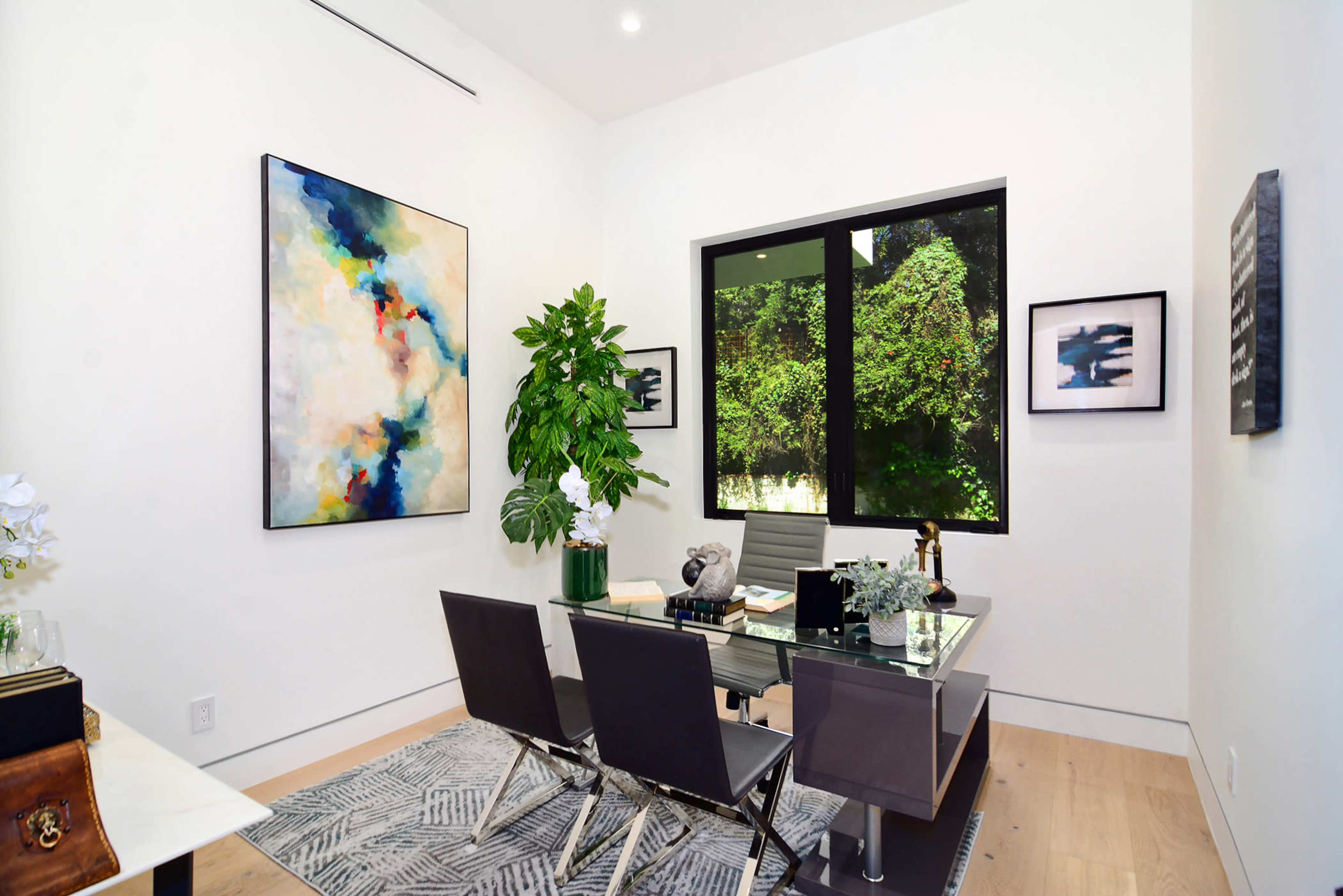 A modern office space features a glass desk, four chairs, a large abstract painting, and a leafy plant, all set against white walls and a window overlooking greenery.