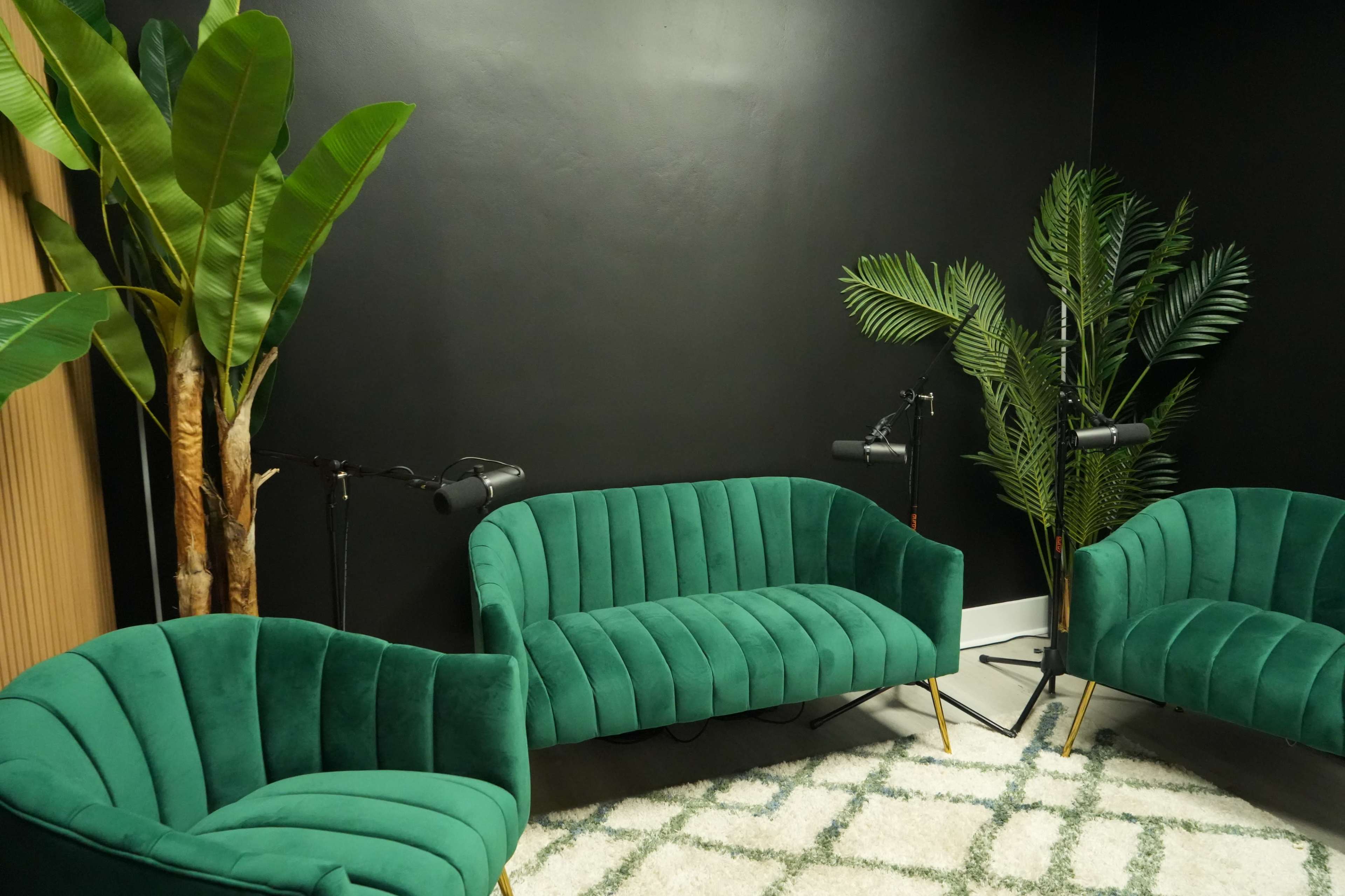 The image shows a cozy seating area with three green velvet chairs, a textured rug, and large potted plants against a dark wall.
