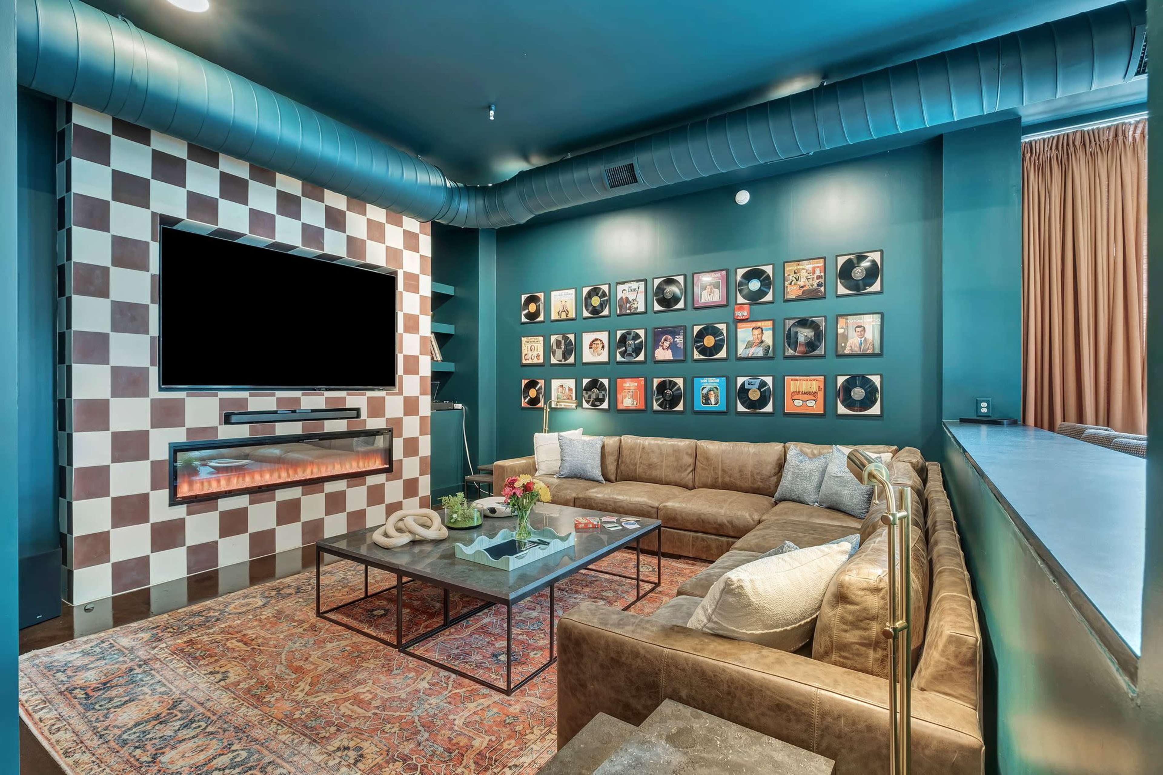 The image depicts a modern living room featuring a brown sectional sofa, a coffee table, a wall adorned with framed records, and a mounted television above a fireplace.