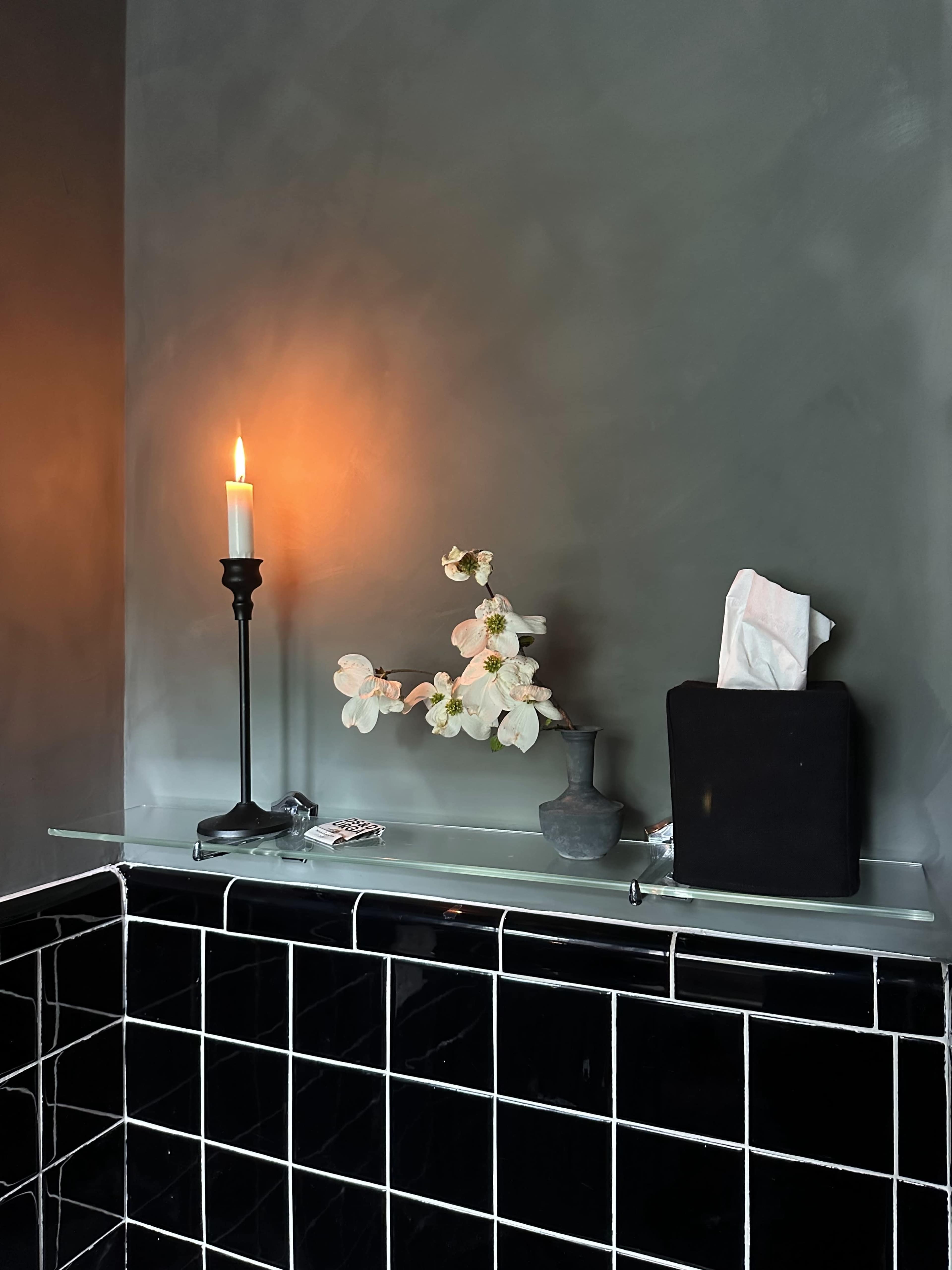 A candle stands on a black stick next to a vase with flowers on a glass shelf above black tiled walls.