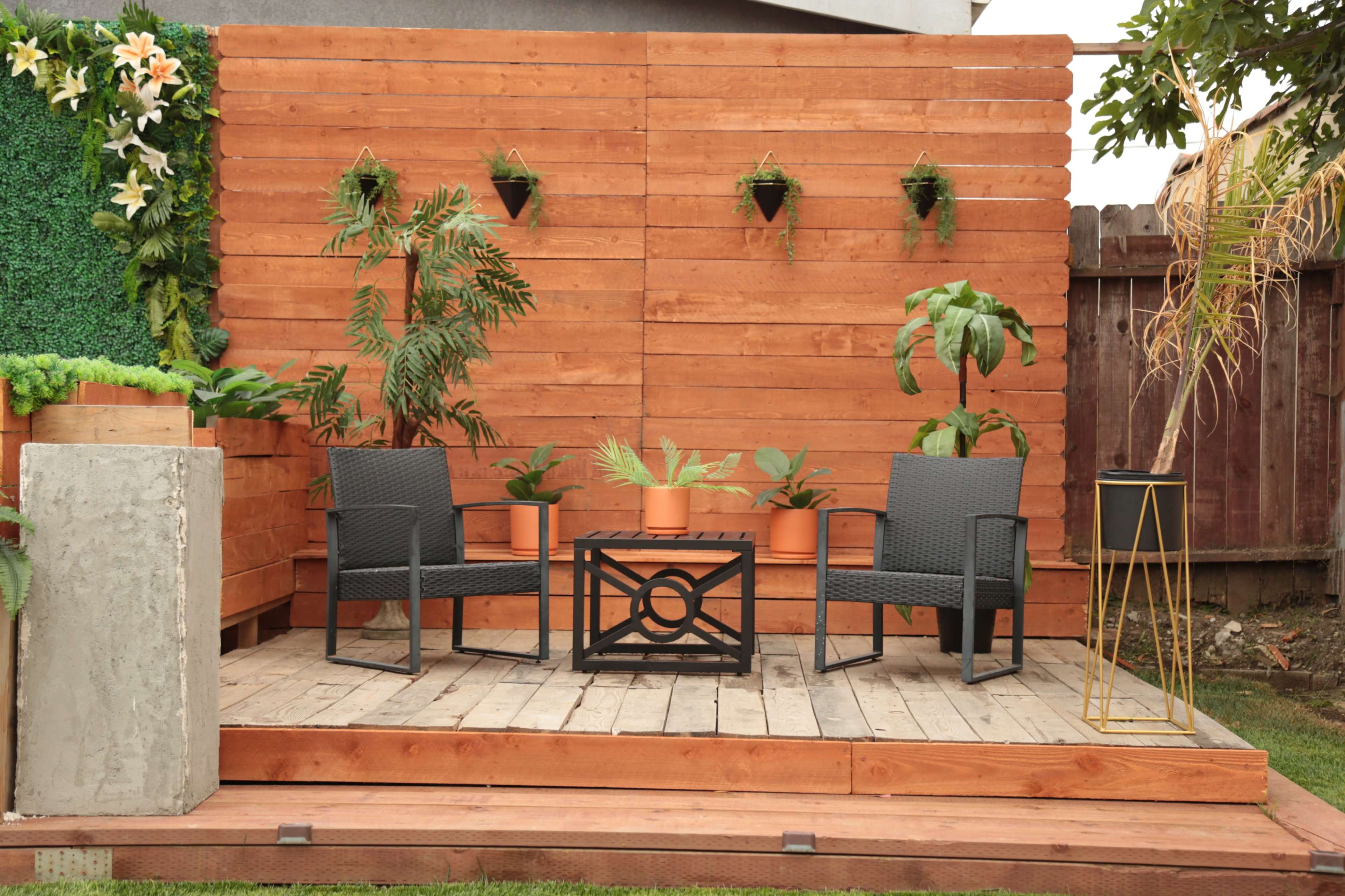 A wooden deck features two chairs and a table surrounded by potted plants against a backdrop of vertical wooden slats.