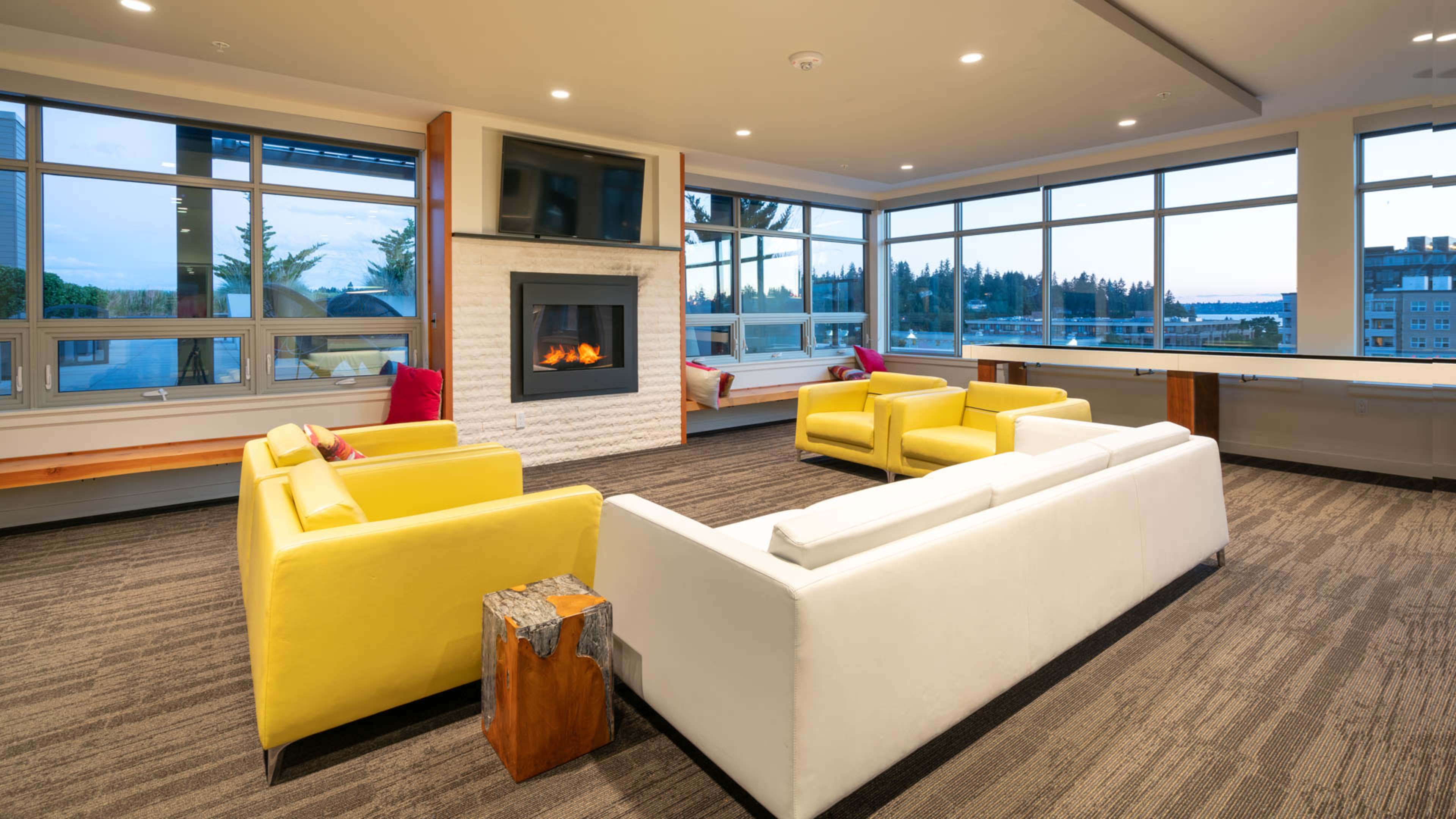 A modern lounge area with two yellow sofas and a white sofa arranged around a fireplace, featuring large windows that provide a view of the outdoors.