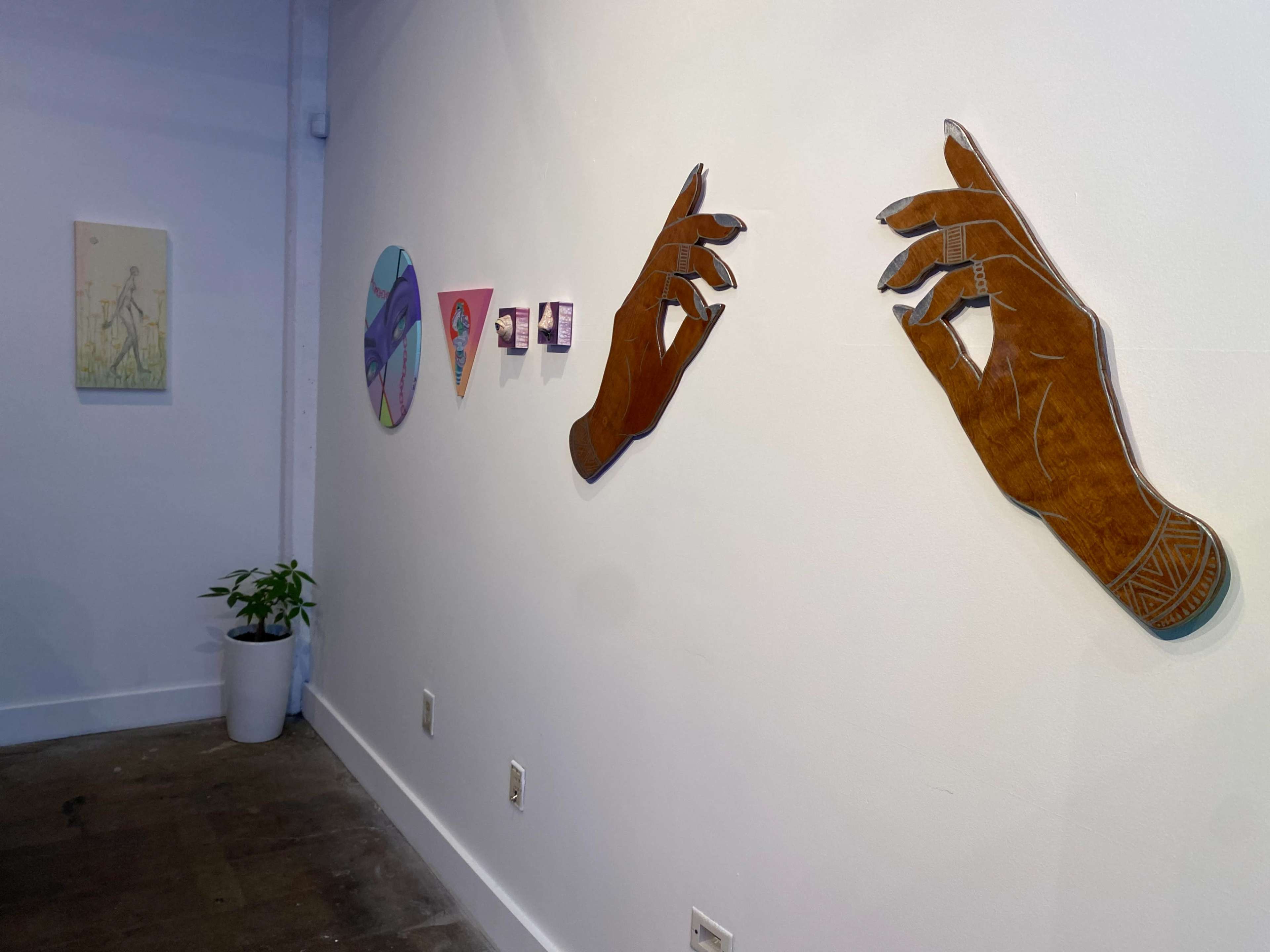 A gallery wall featuring various art pieces, including wooden hand sculptures, framed images, and a potted plant.