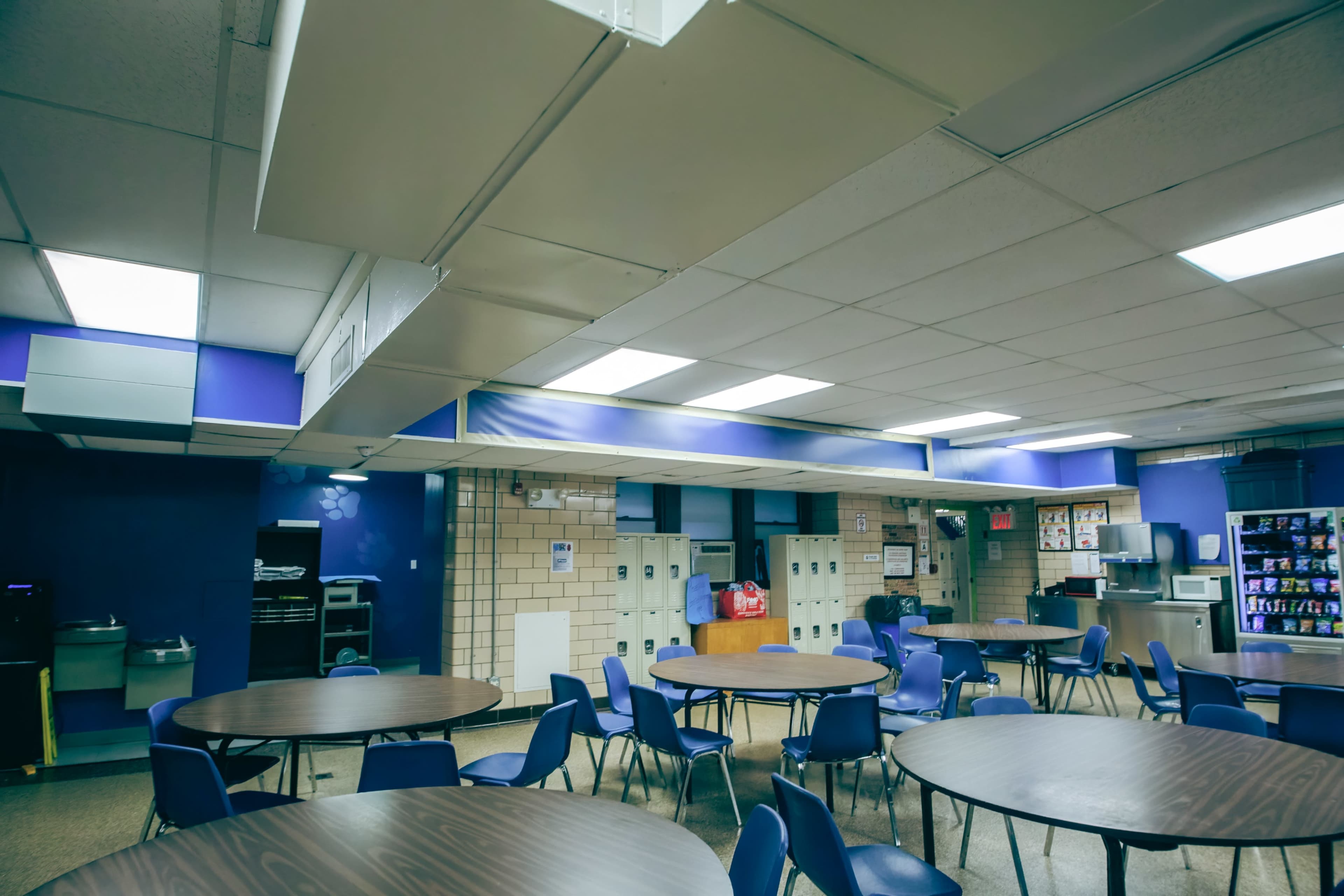 A cafeteria with several circular tables and blue chairs, along with lockers and vending machines in the background.