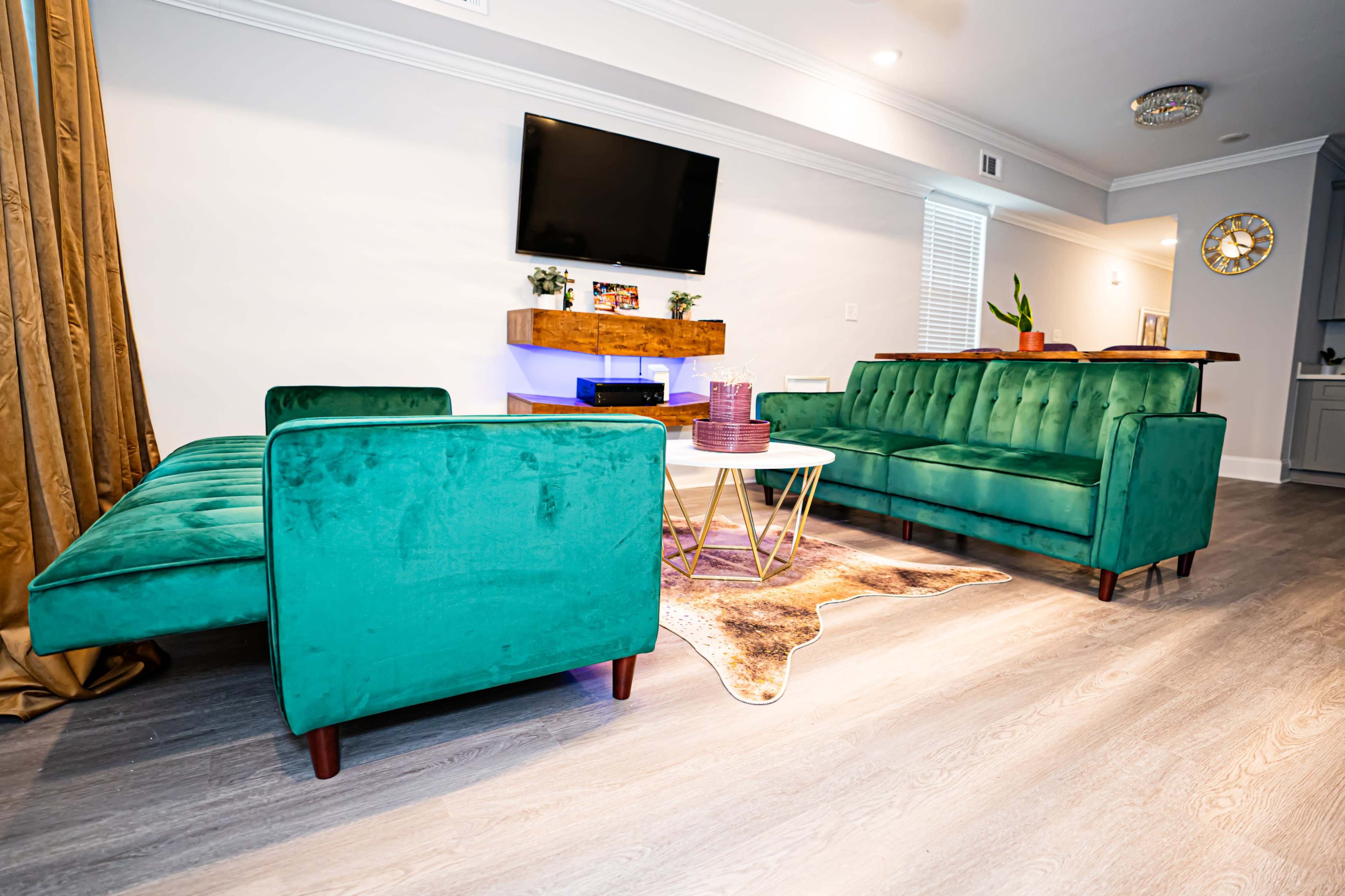 A modern living room features green velvet sofas arranged around a central coffee table, with a wall-mounted TV above a wooden shelf.