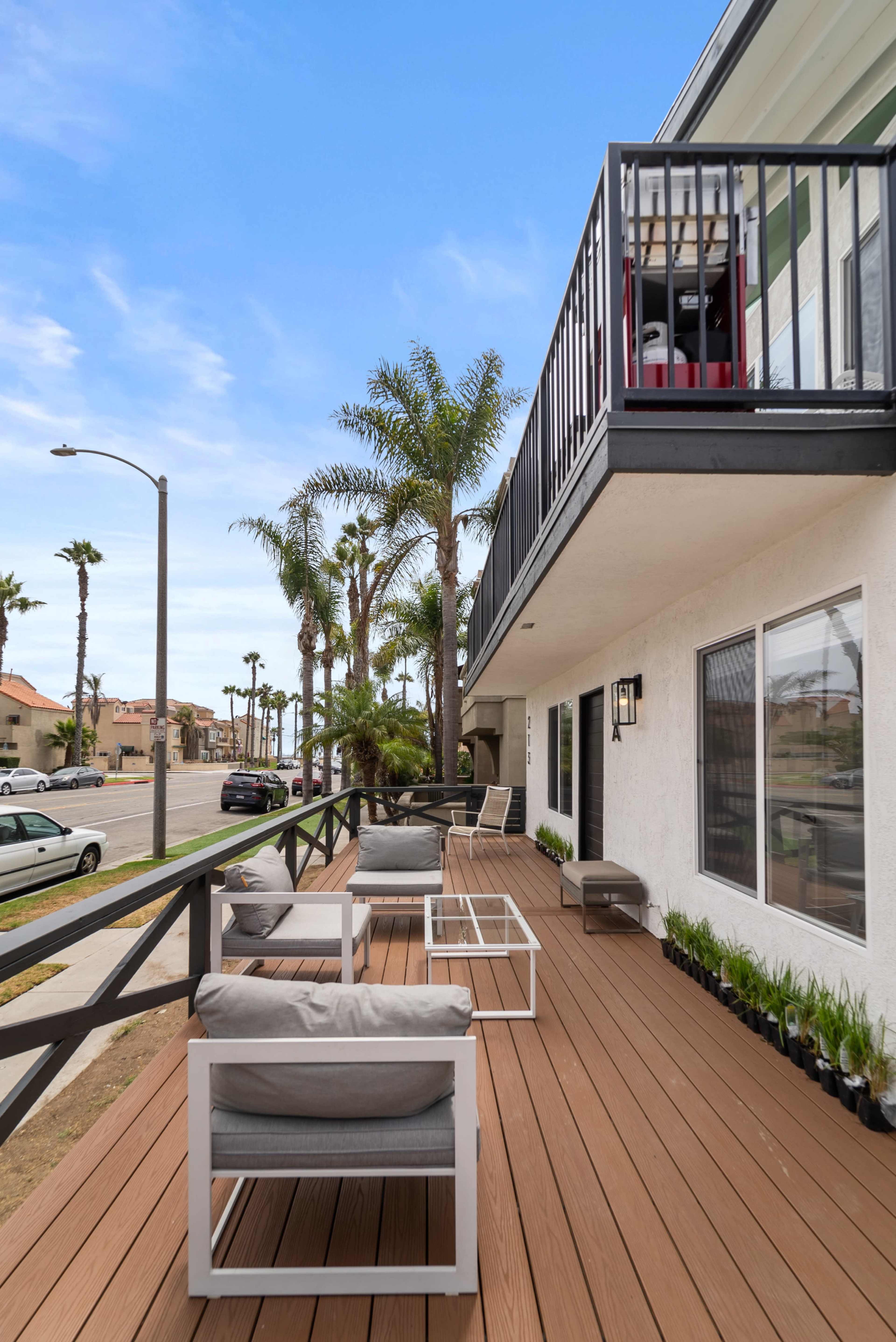 A wooden deck with modern furniture is located on the second floor of a building, surrounded by palm trees and facing a street.