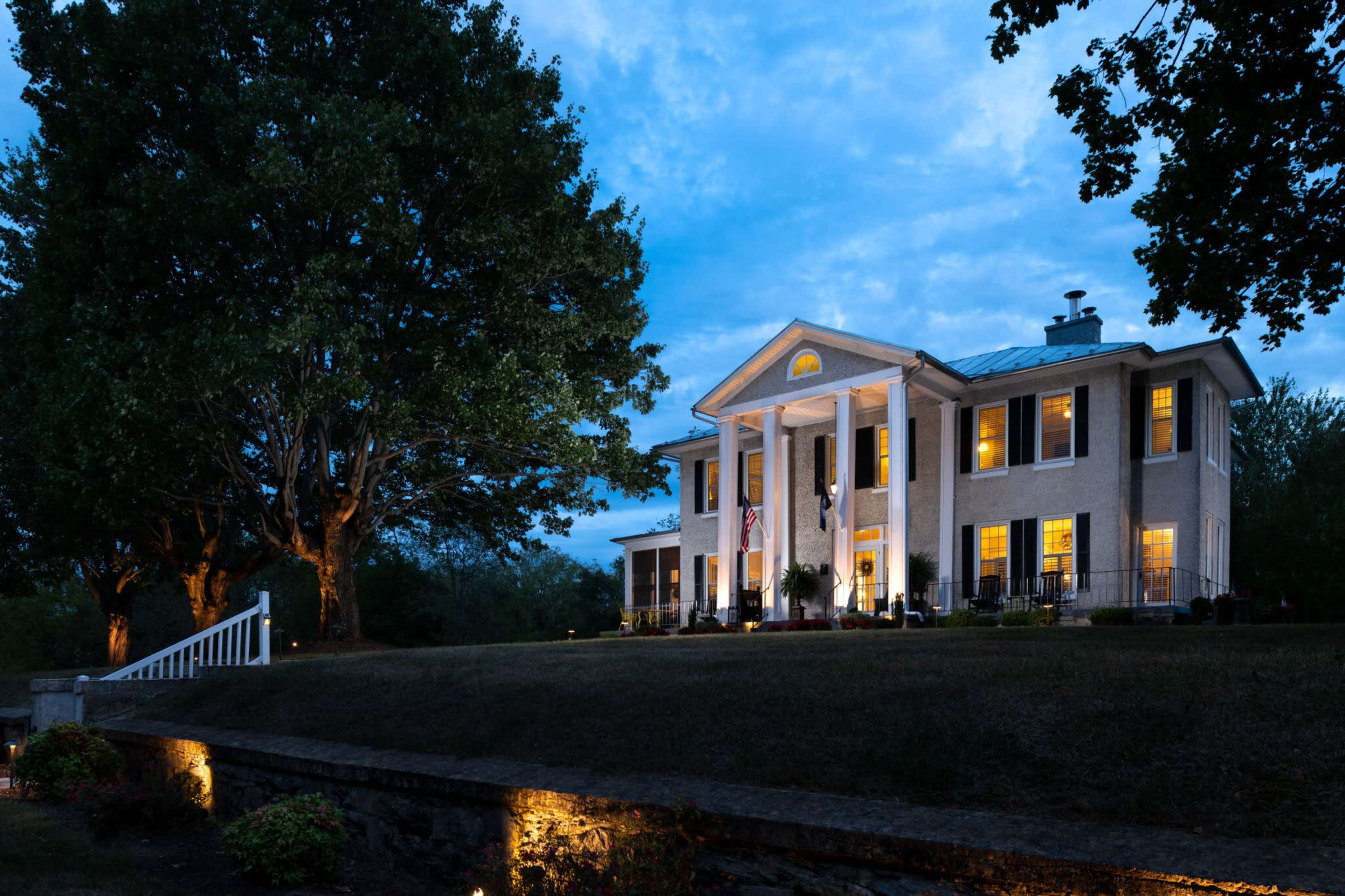 A large, two-story house with white columns is illuminated in the evening light, surrounded by trees and landscaped shrubs.