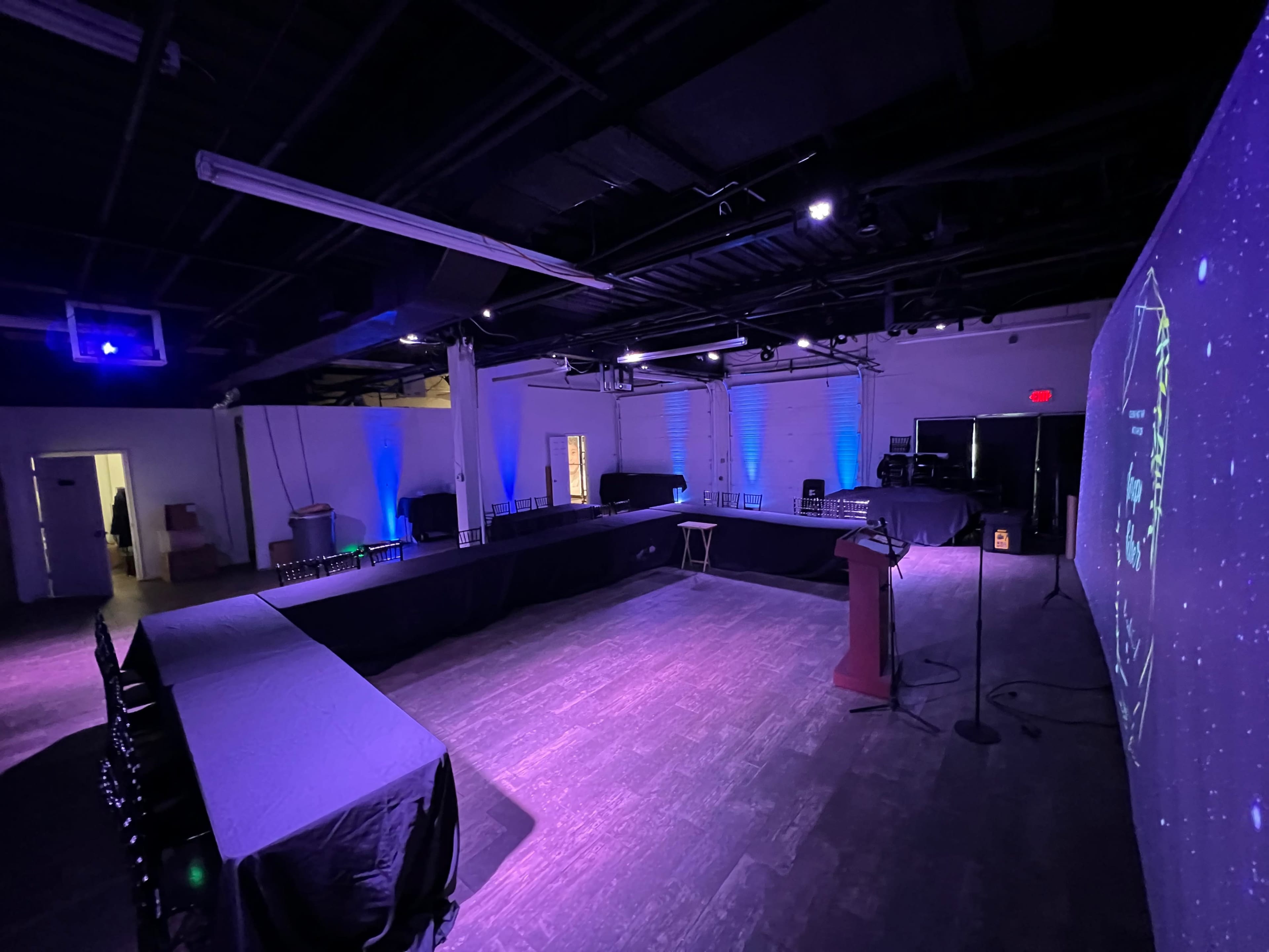 An empty event space with a rectangular arrangement of tables, chairs, and a podium, illuminated by blue lighting and featuring a projection on one wall.