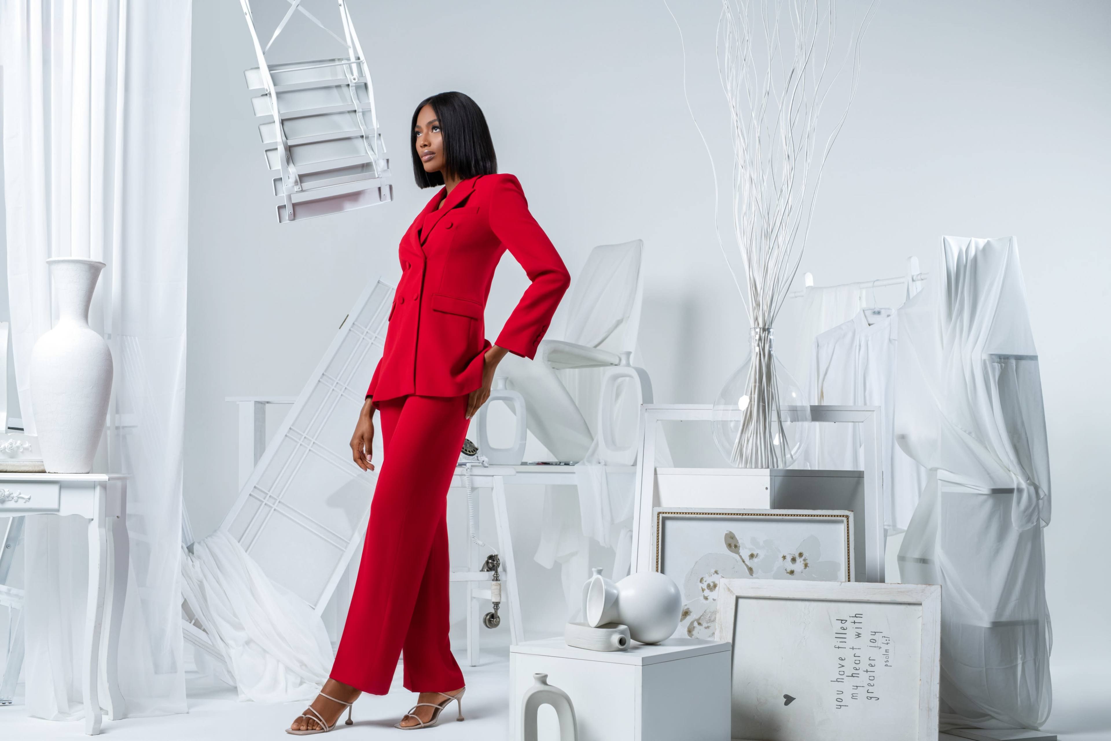A model poses in a red suit among a minimalist white backdrop with various pieces of furniture and decor.