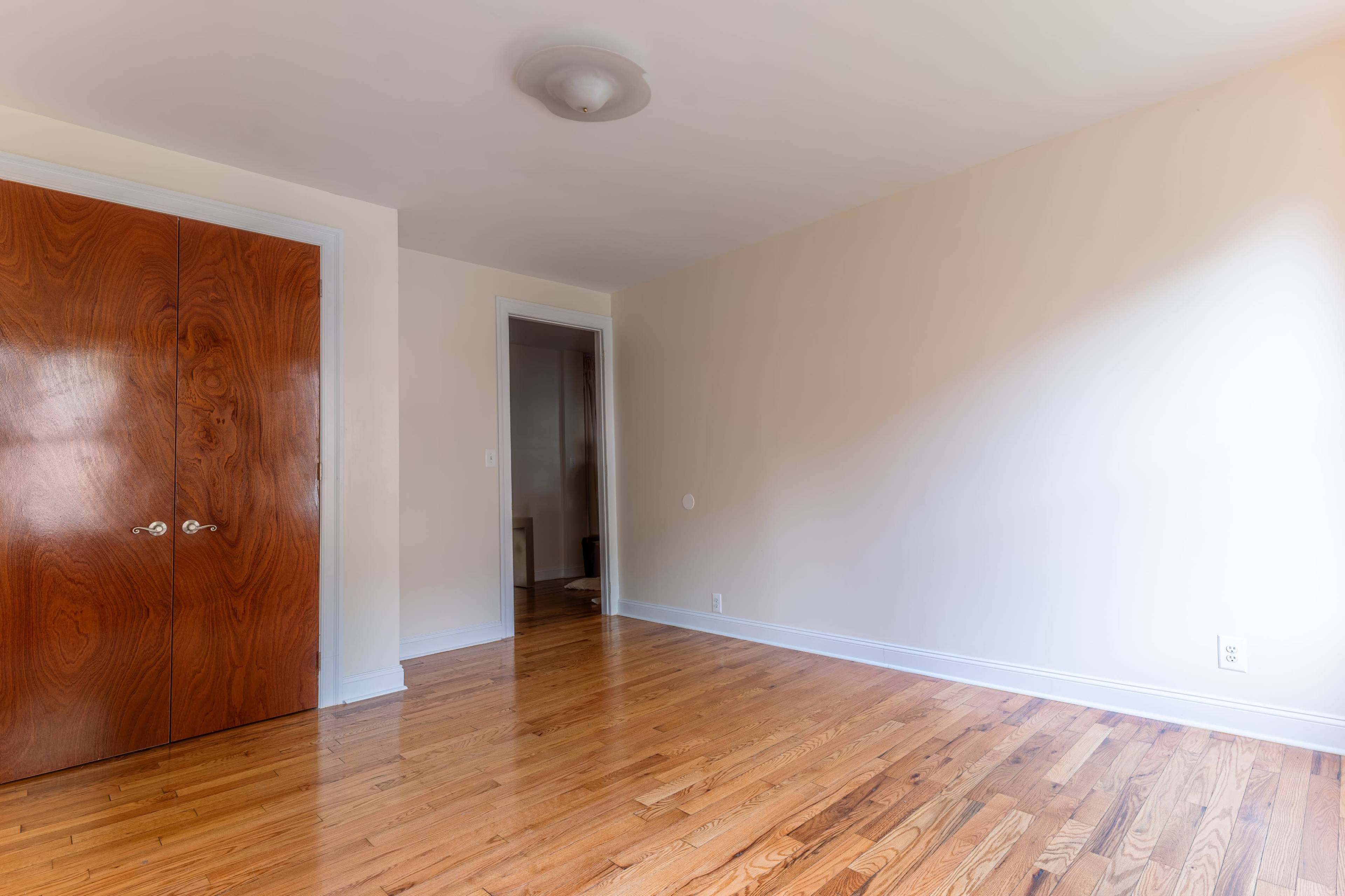 The image shows a vacant room with hardwood floors, a ceiling light fixture, and a closet with wooden doors.
