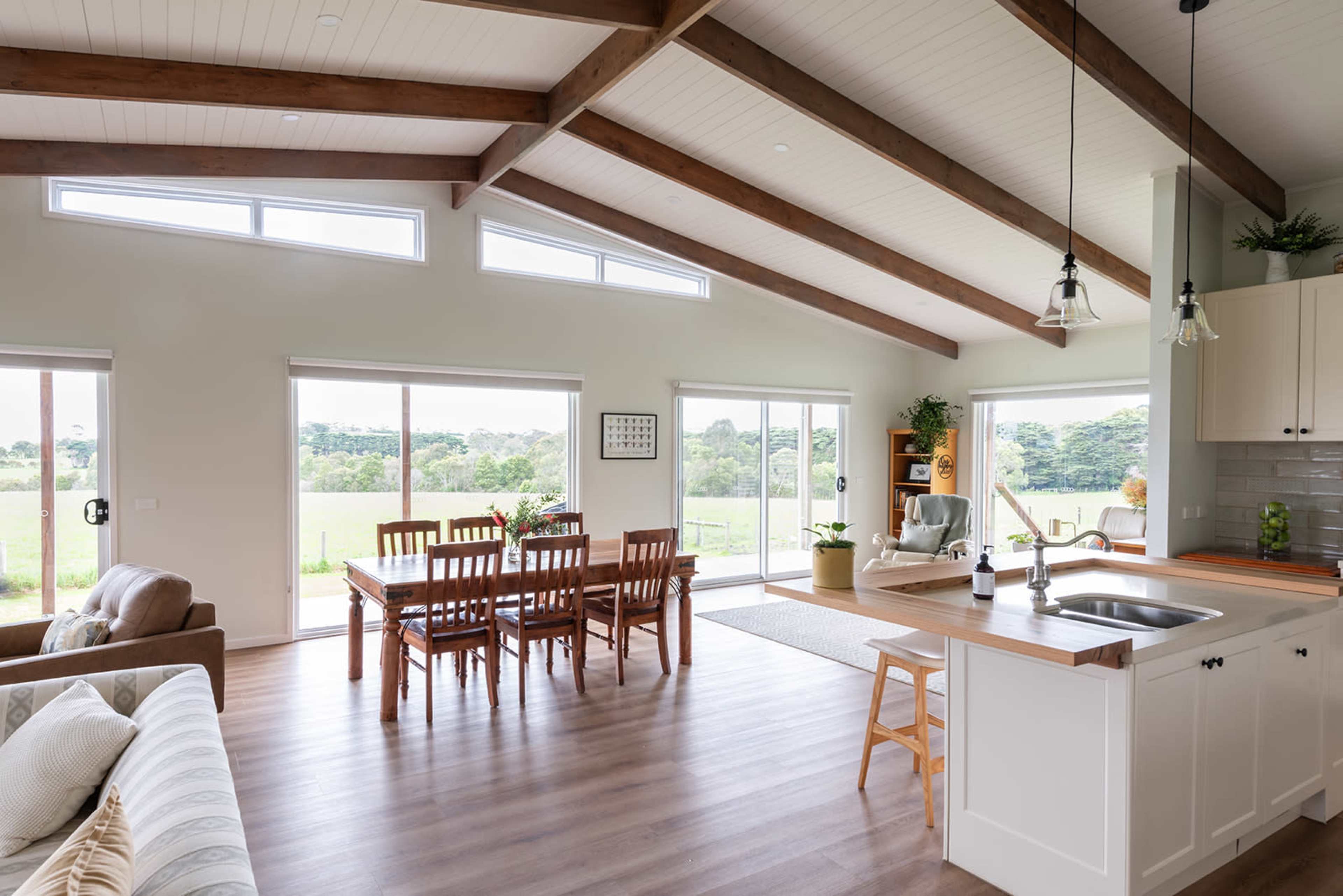 The open-concept living area features a dining table, kitchen island, and large windows overlooking a green landscape.