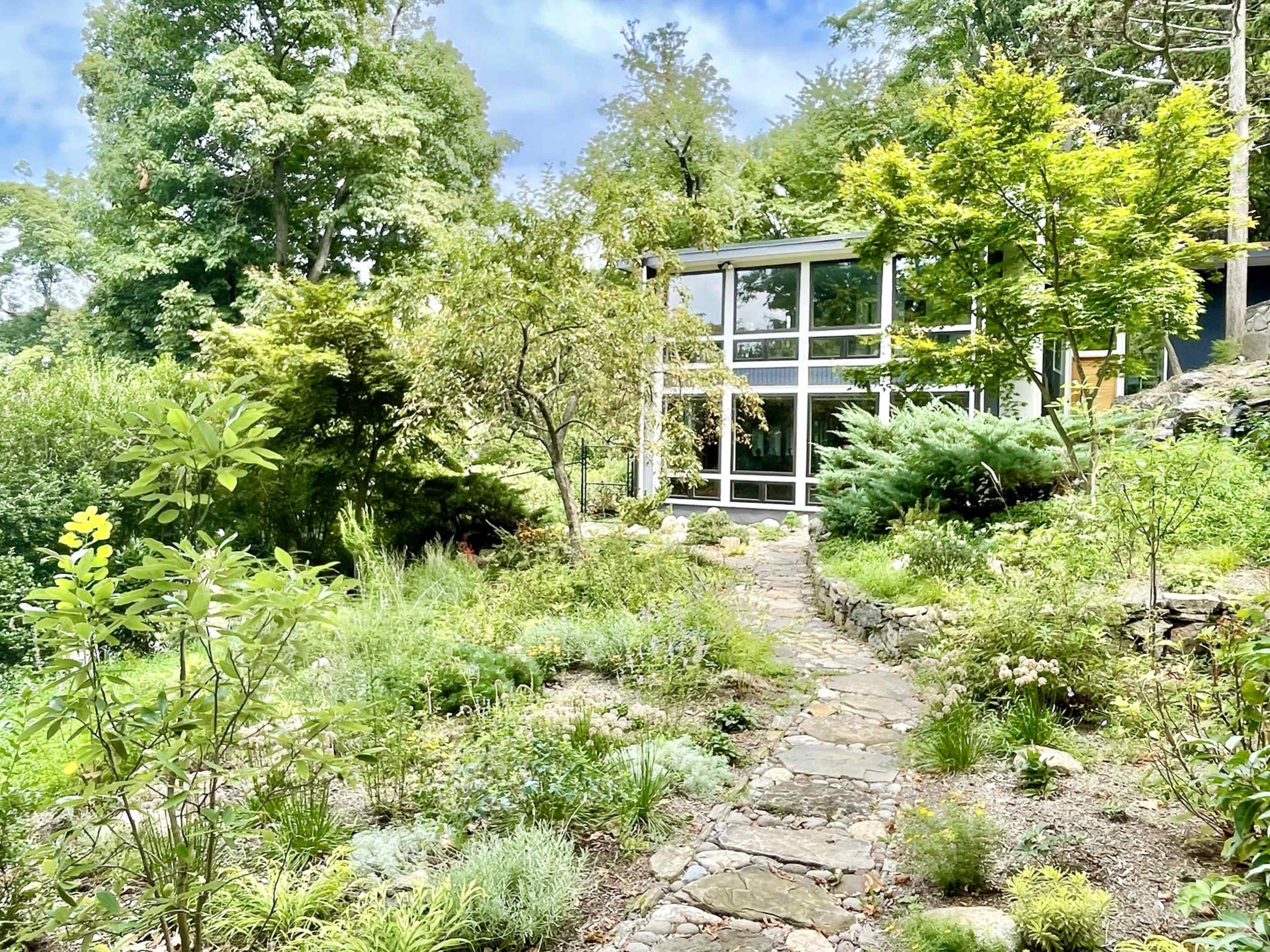 A stone pathway winds through a lush garden leading to a modern glass-fronted house surrounded by greenery.
